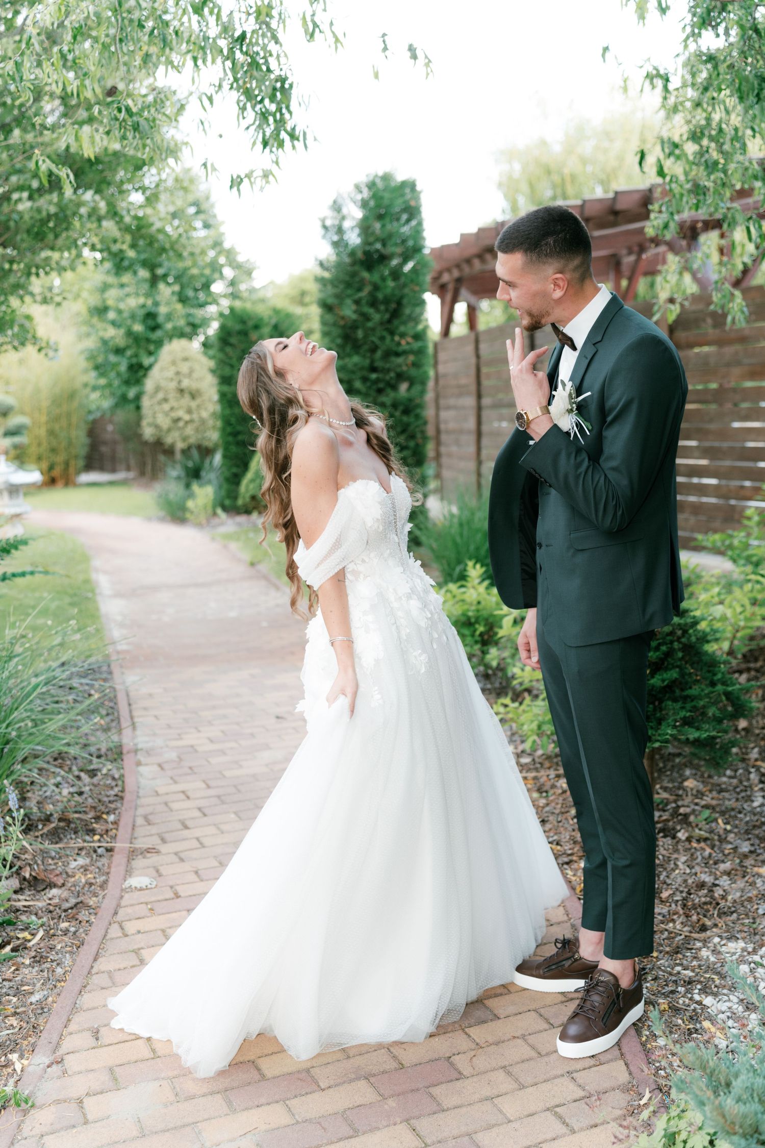 Bride in off-shoulder floral gown laughing joyfully with groom on brick garden pathway. Mallorca wedding photographer and filmmaker.