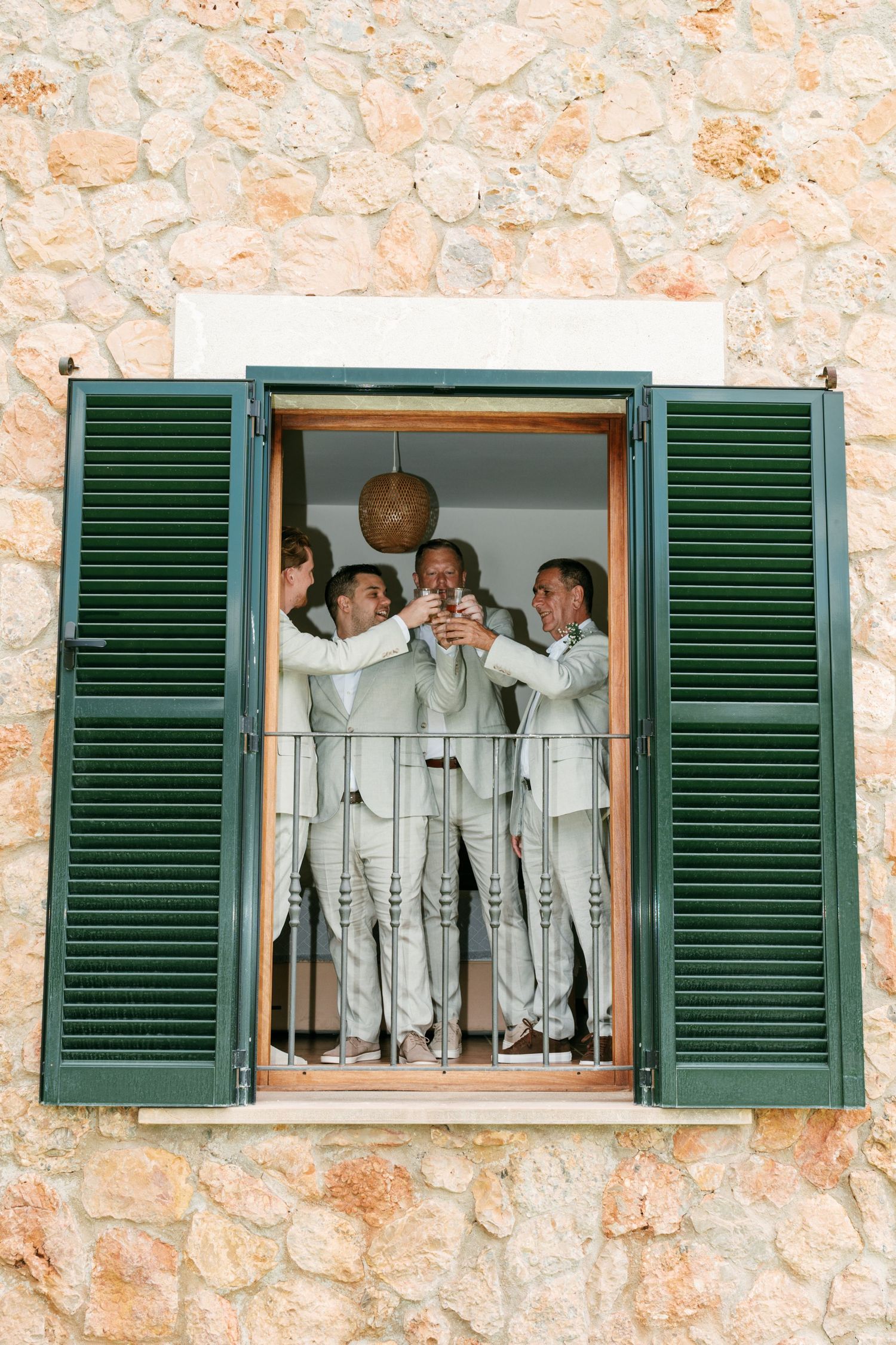 Groomsmen in sage suits toasting drinks together in an open green-shuttered window of a rustic stone building. Mallorca wedding photographer