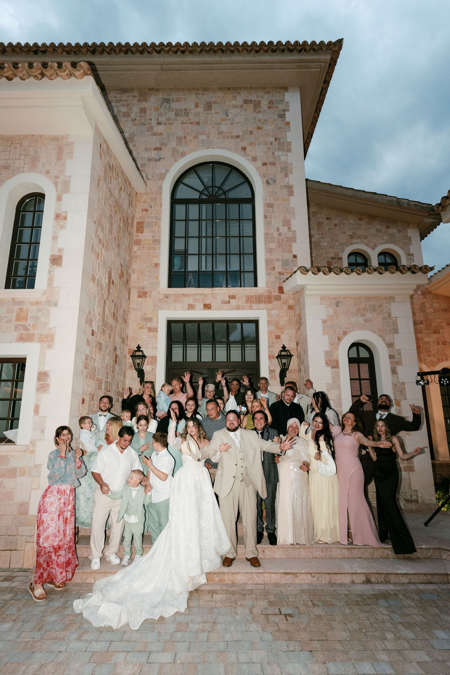 Wedding party and guests pose on steps of elegant stone mansion for group celebration photo.
