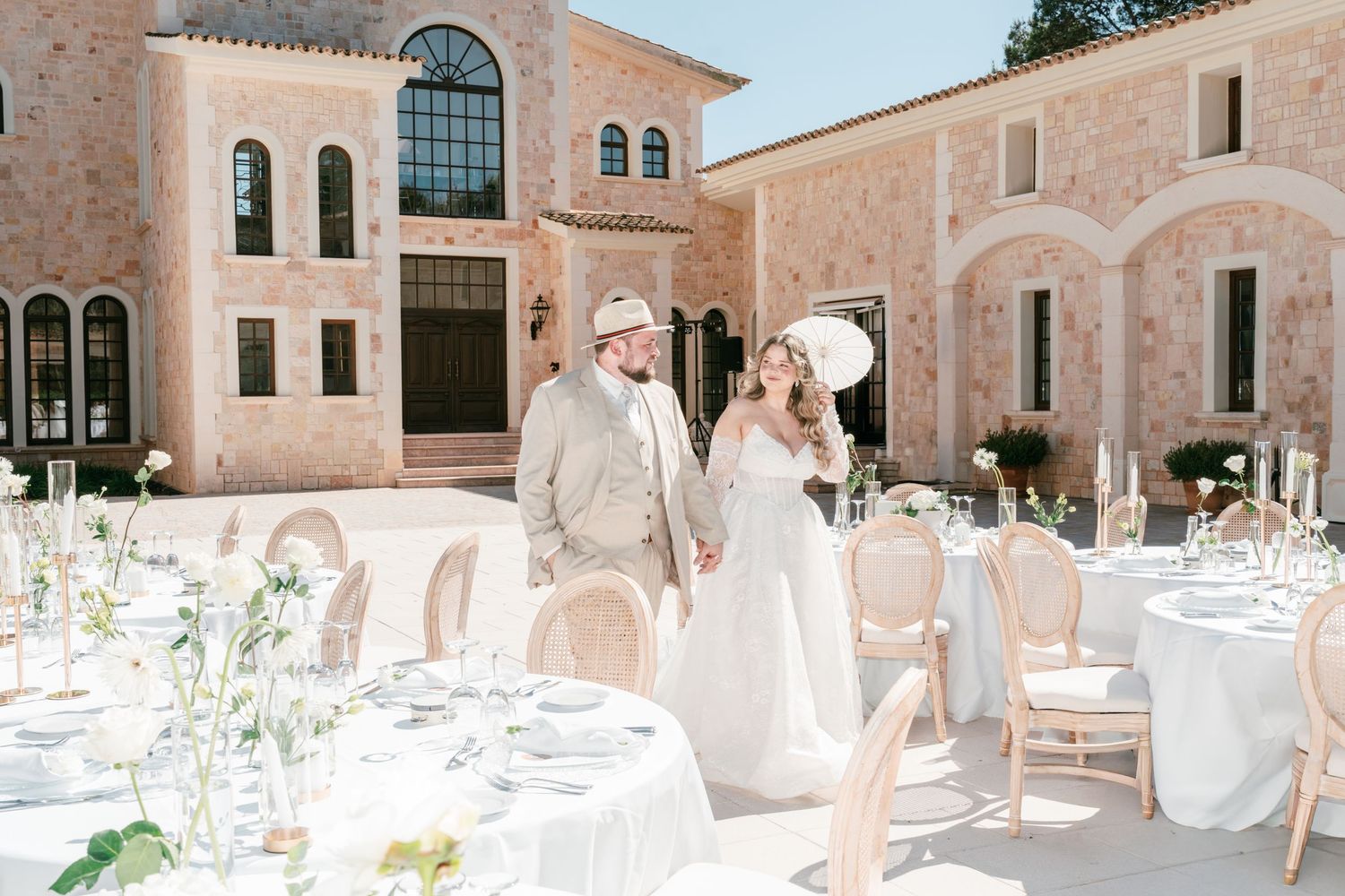 Bride and groom hold hands at elegant outdoor wedding reception with white tablecloths at a pink Mediterranean villa in Mallorca.