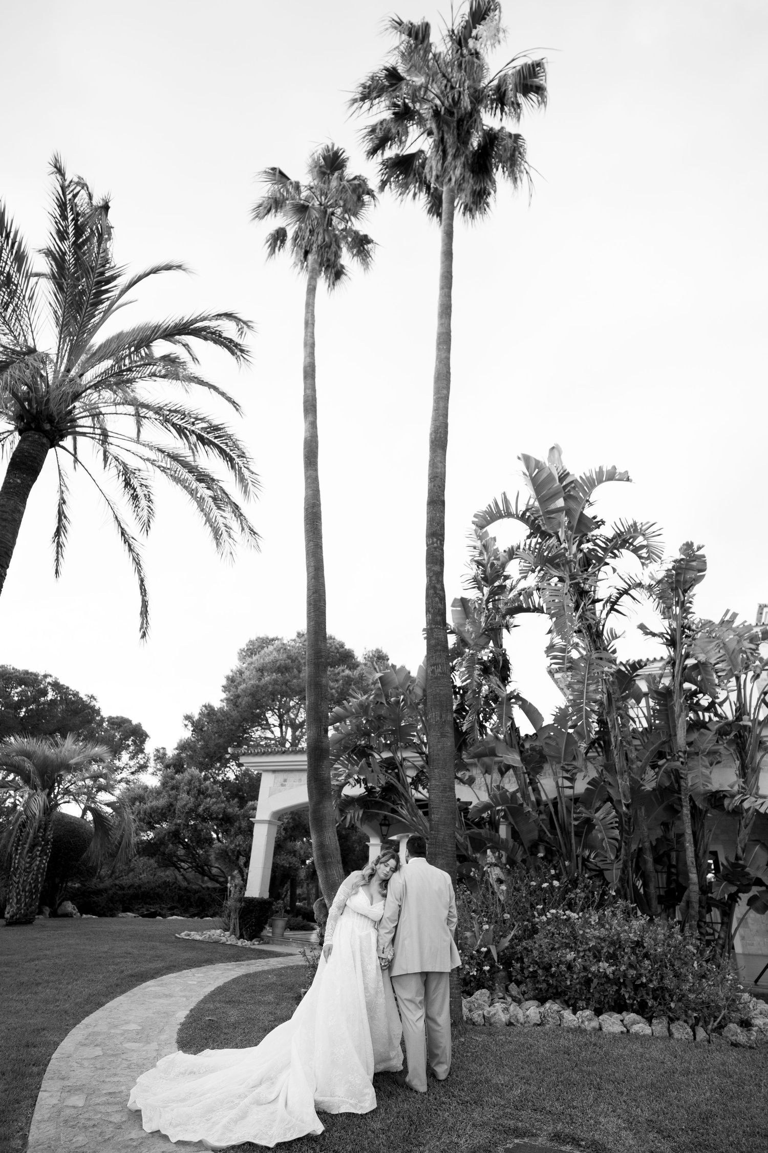Black and white photo of a bride and groom embracing in a tropical garden with tall palm trees.