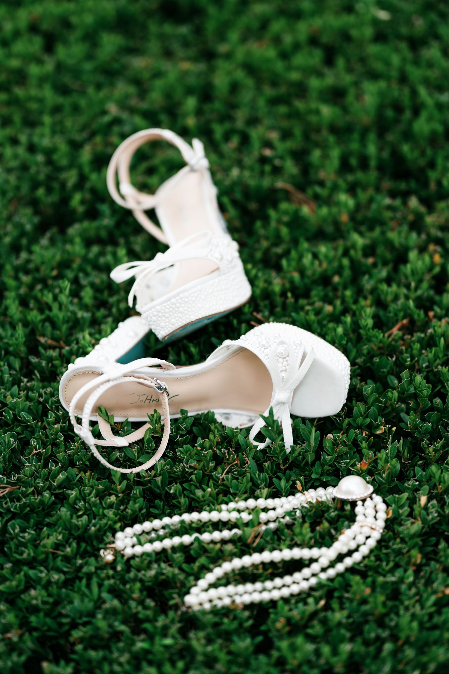 White platform bridal sandals with ankle straps and lace bows rest on green grass beside a pearl necklace.