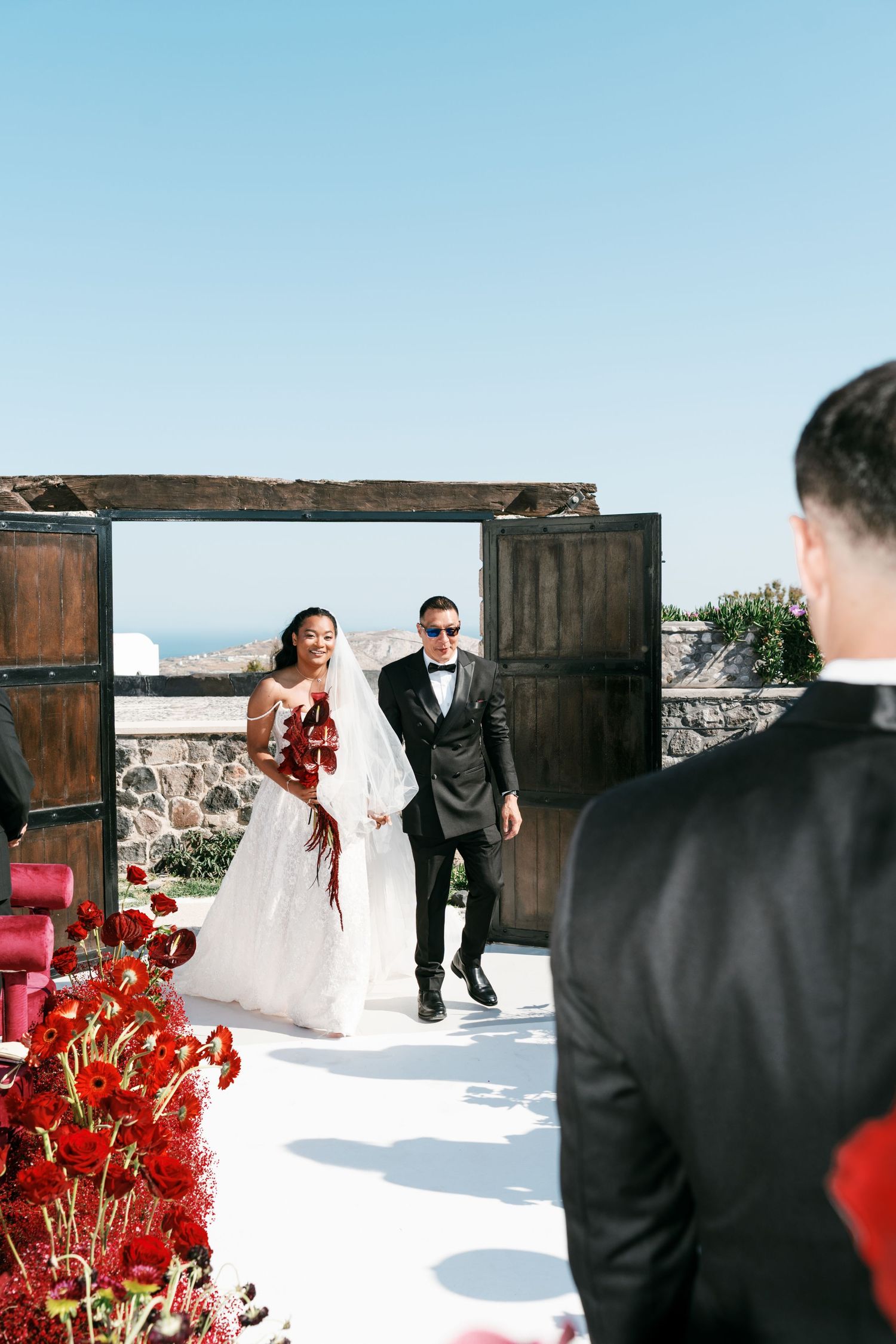 Bride in white gown with red bouquet walks down aisle with escort toward groom at outdoor wedding ceremony.