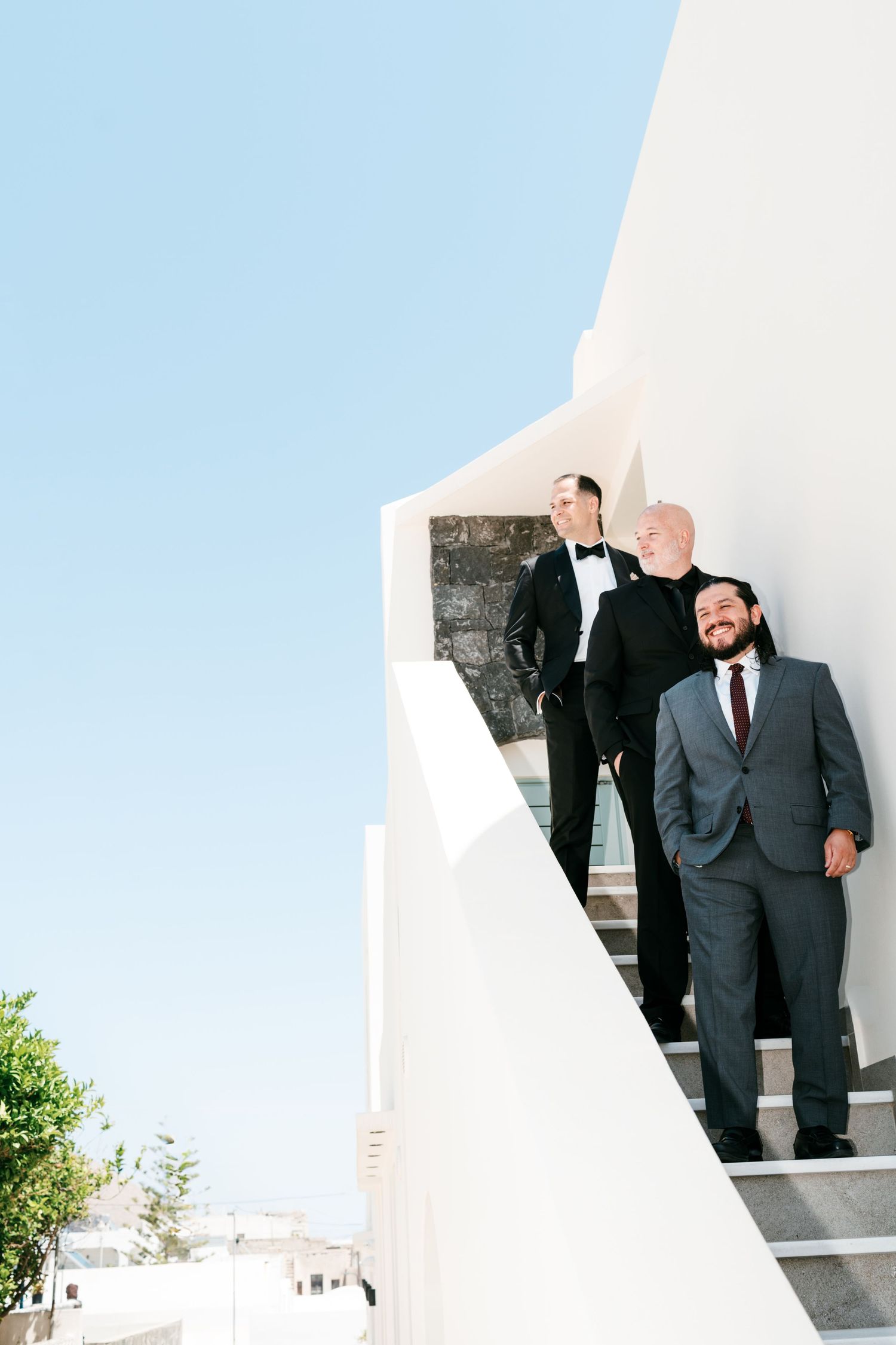 Three groomsmen in formal suits descend white Mediterranean stairs under a clear blue sky.