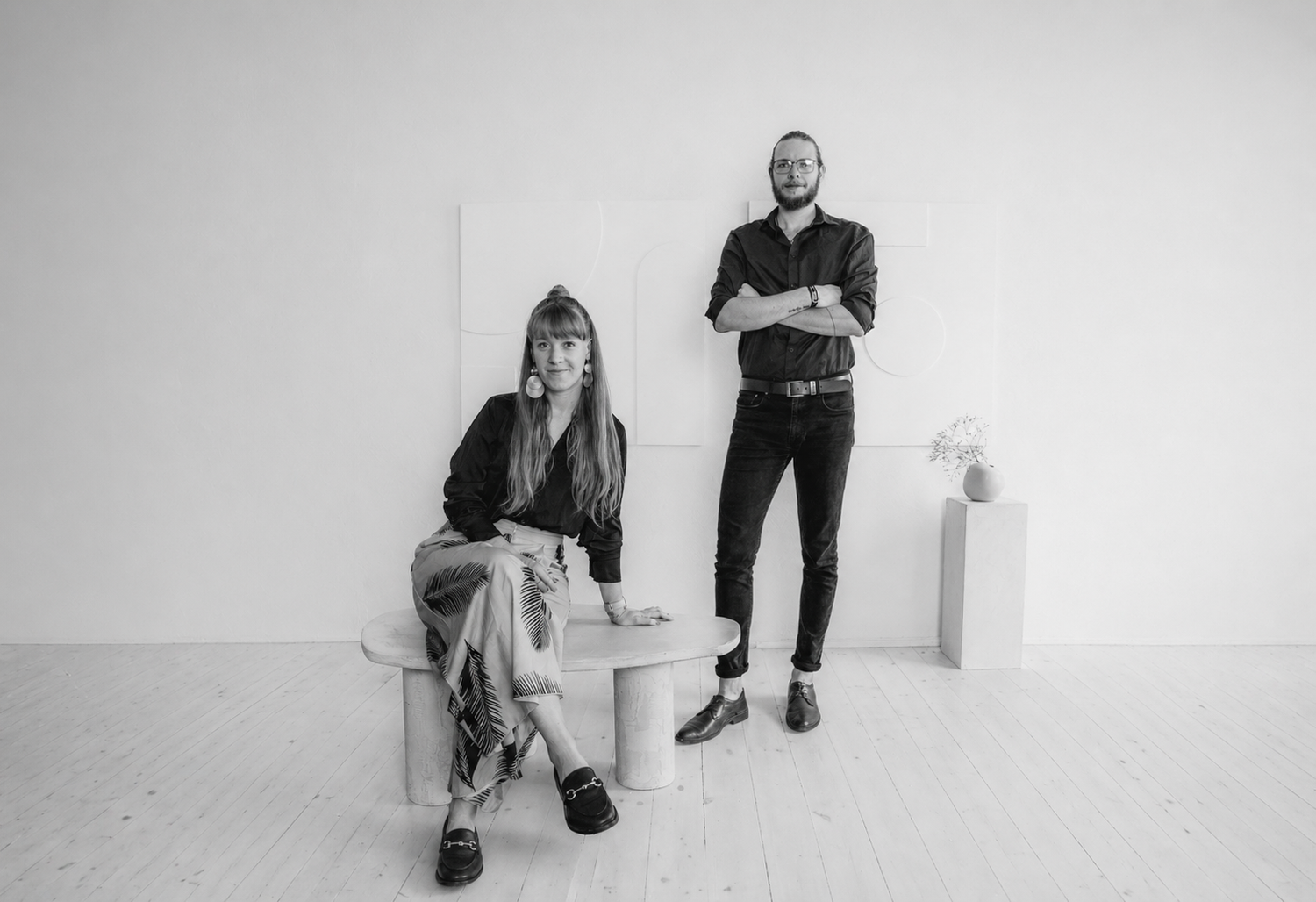 Black and white photo of a woman sitting and a bearded man standing in a minimalist white studio setting.