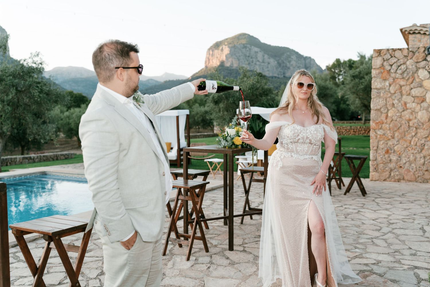 Couple toasting with red wine glasses outdoors by a pool with mountain views at a Mediterranean villa . Mallorca wedding photographer.