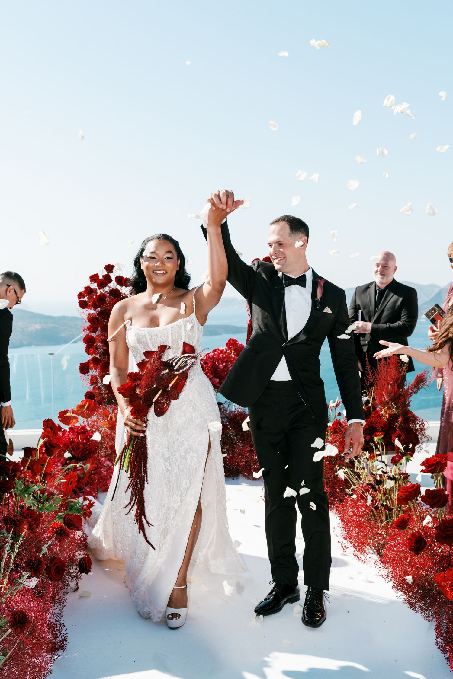 Joyful newlyweds walk down the aisle amid falling petals, surrounded by red roses, with a stunning ocean backdrop.