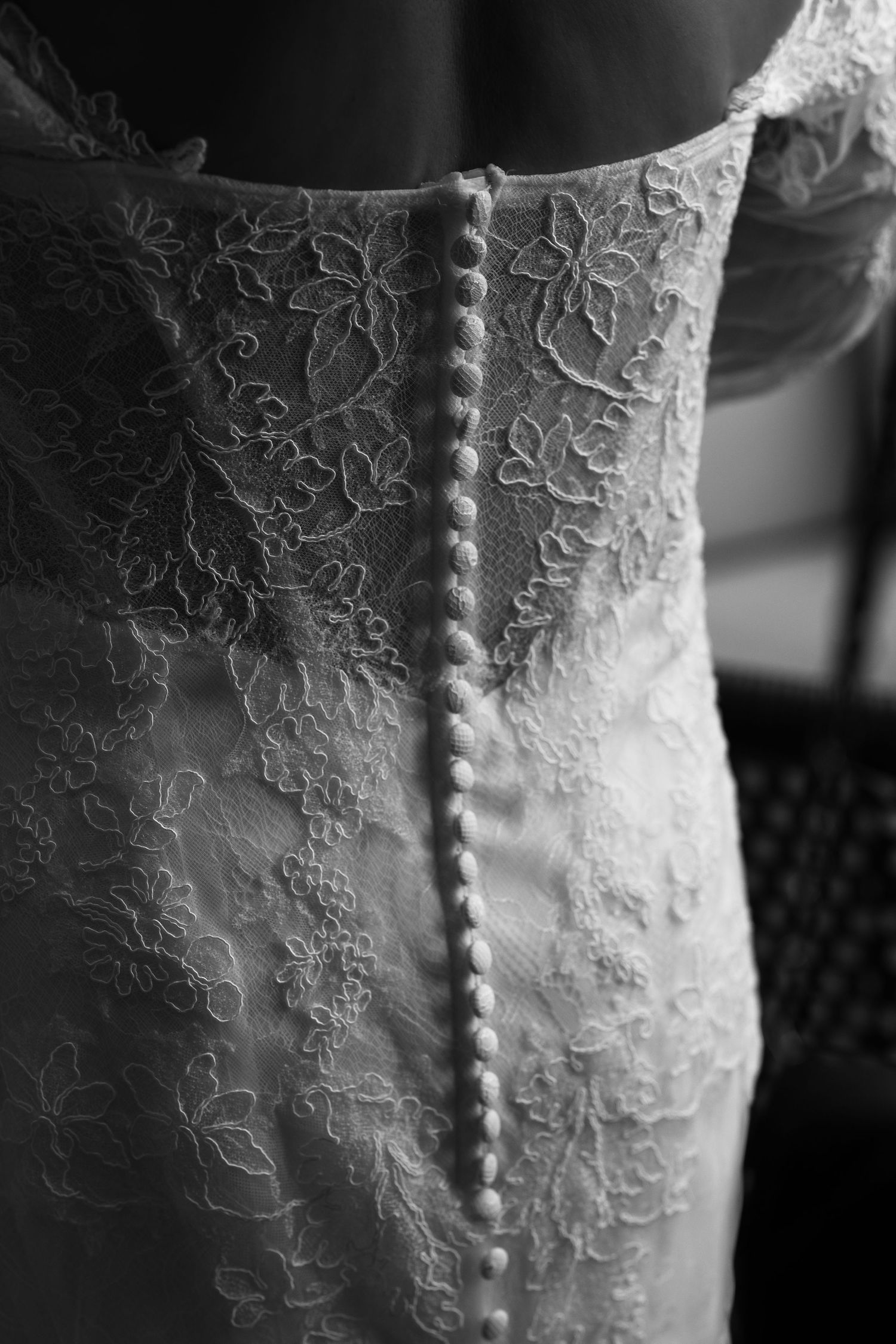 Black and white close-up of a lace wedding dress back with a row of small fabric-covered buttons and delicate floral lace detail.