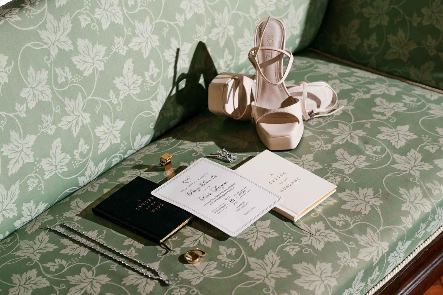 Blush bridal heels, wedding invitation suite, black ring box and gold rings arranged on a sage green floral vintage sofa.