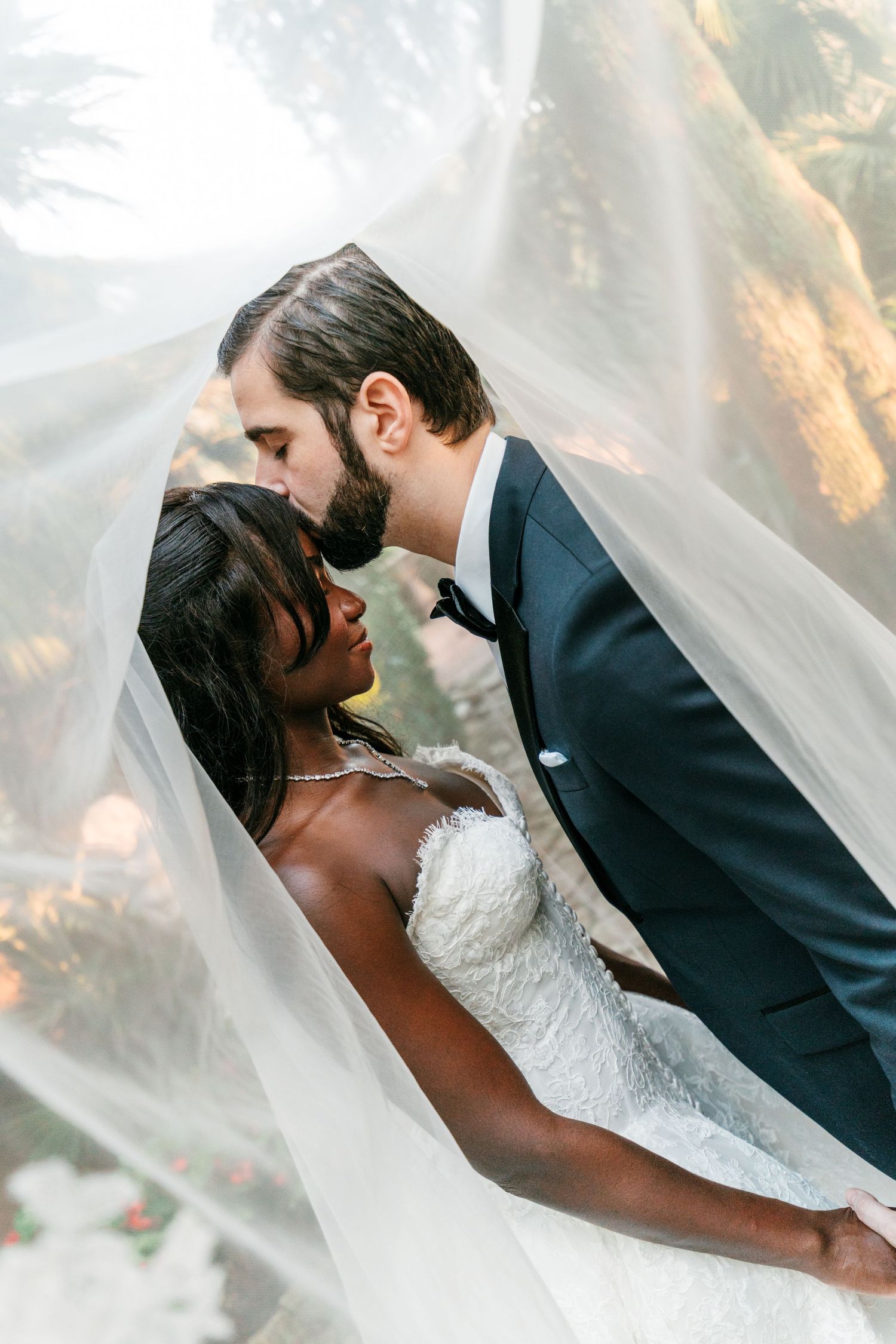 Groom in a navy tuxedo and bow tie kisses his bride's forehead as a sheer wedding veil drapes over them outdoors.