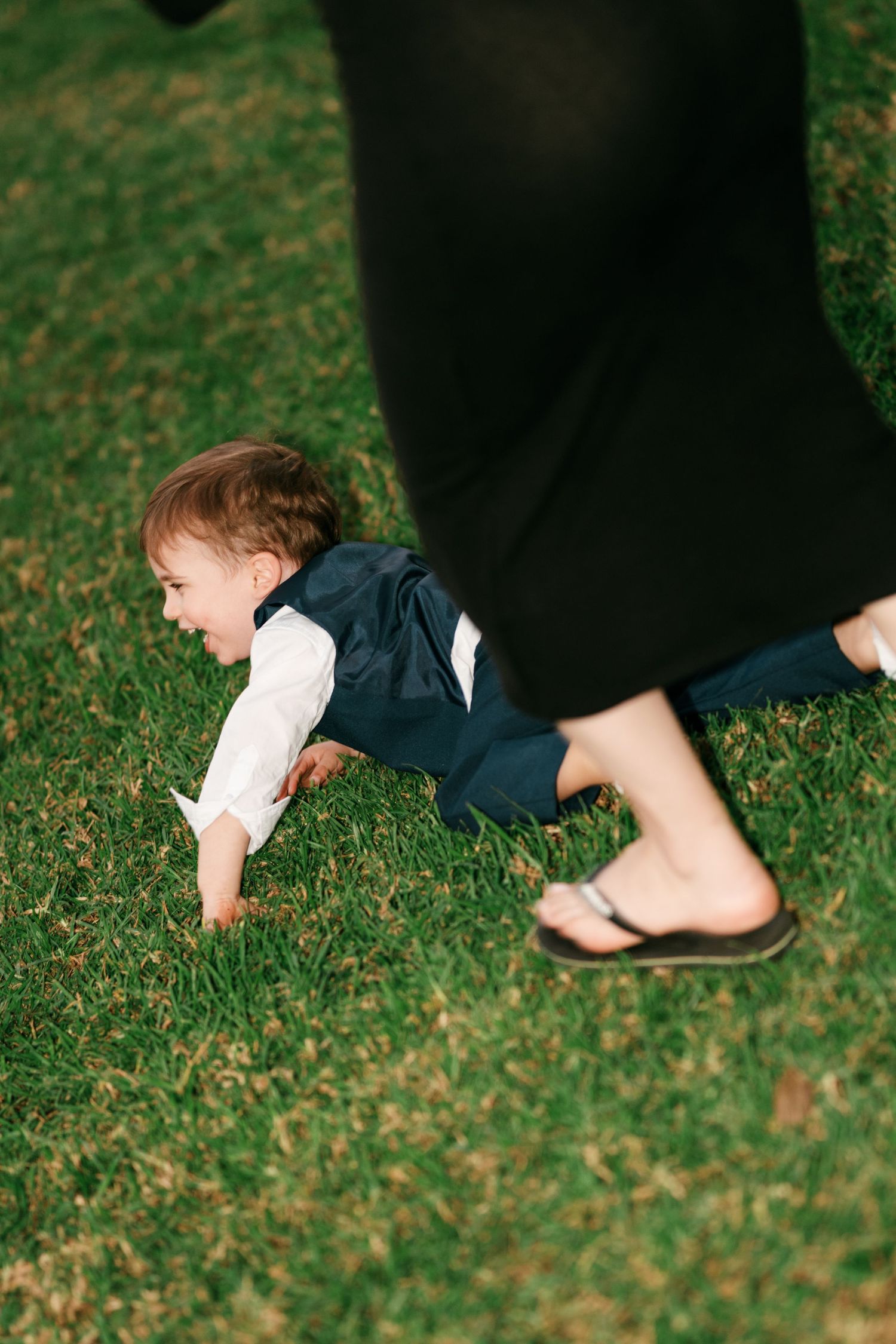 Smiling toddler boy in a navy vest and white shirt crawls on green grass as a person in a long black dress and flip-flops walks past.
