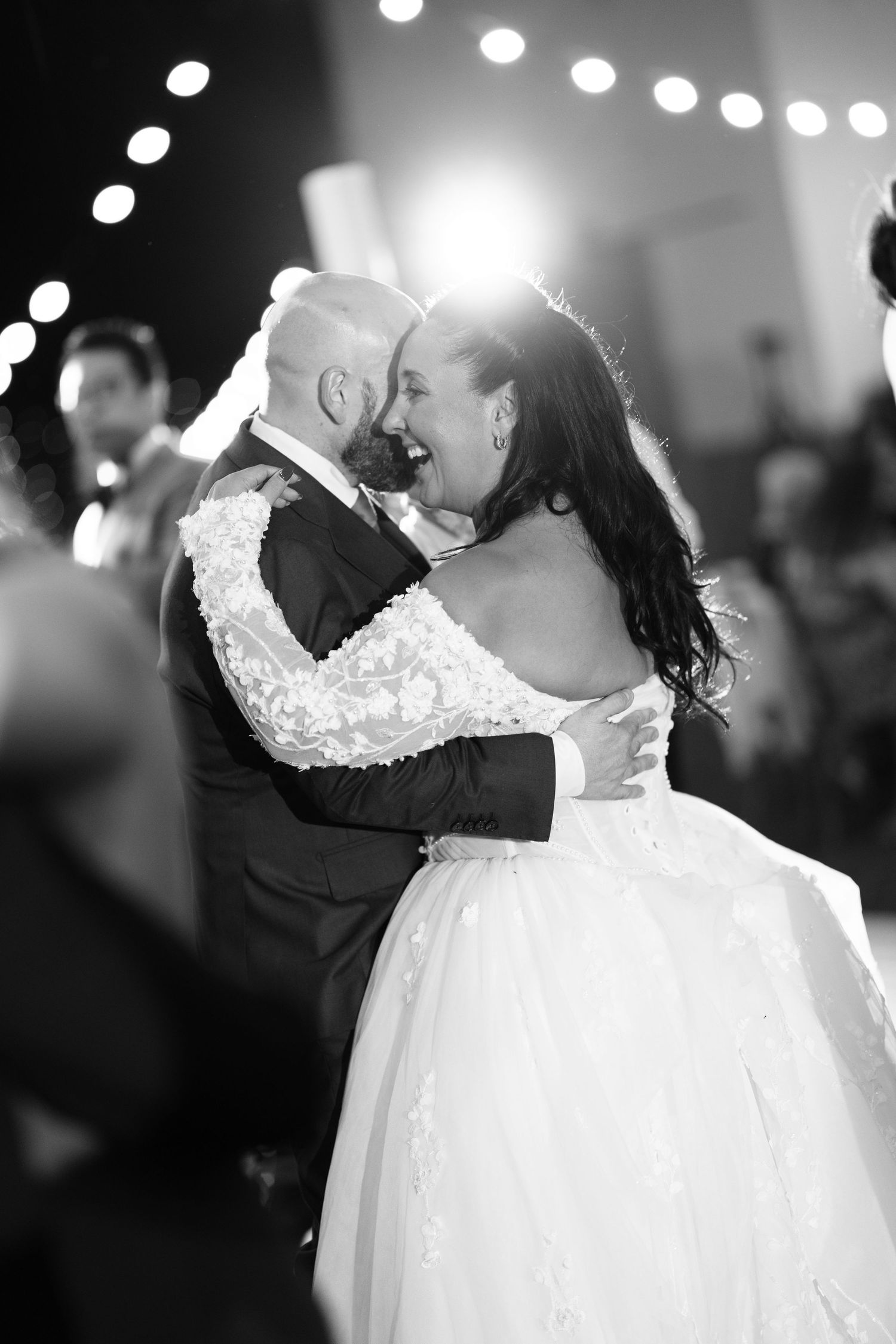 Black and white photo of a bride in a lace off-shoulder wedding gown laughing as she dances closely with her groom during a reception.