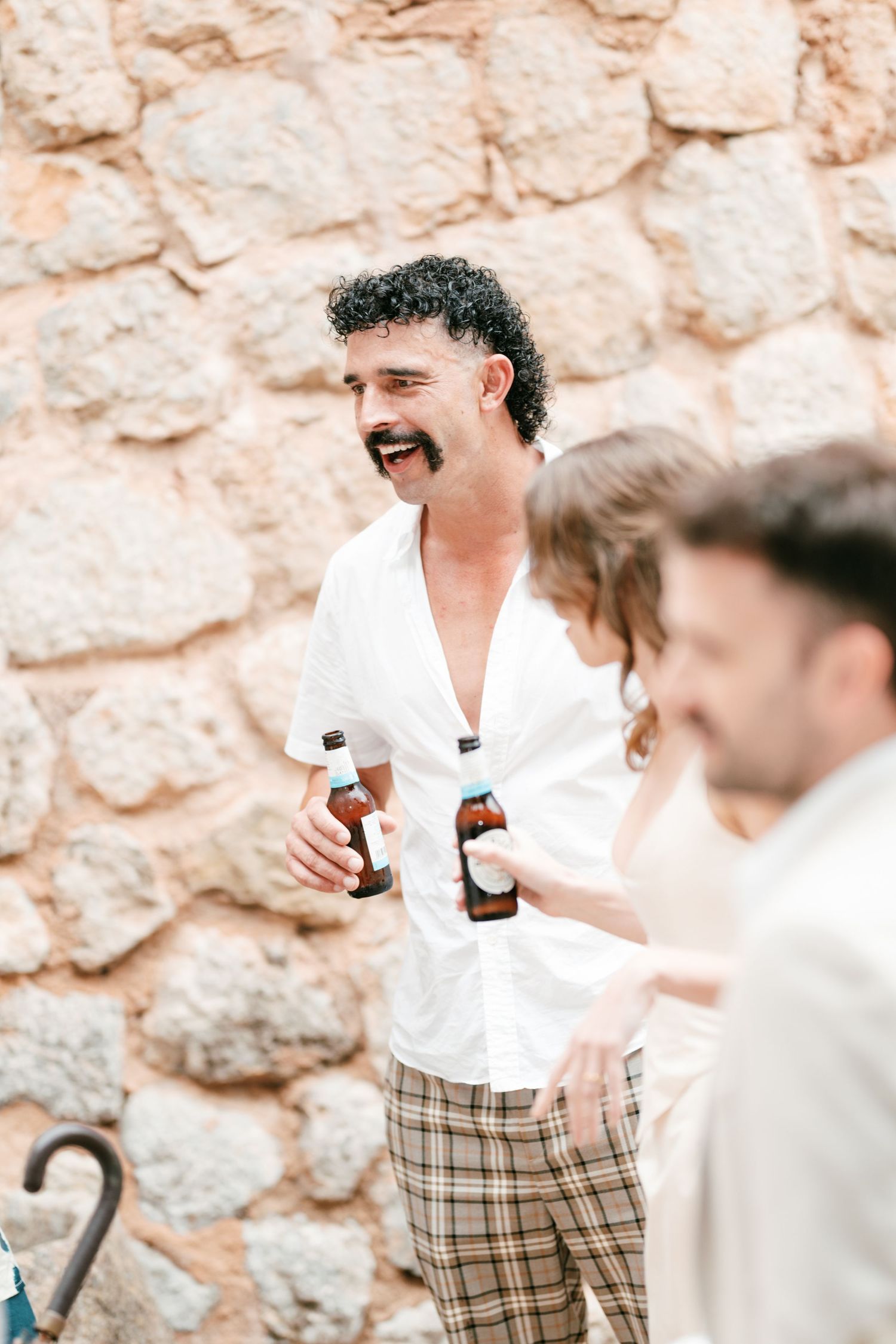 Smiling man with curly hair and a mustache holds two beer bottles beside guests against a rustic stone wall at an outdoor celebration.