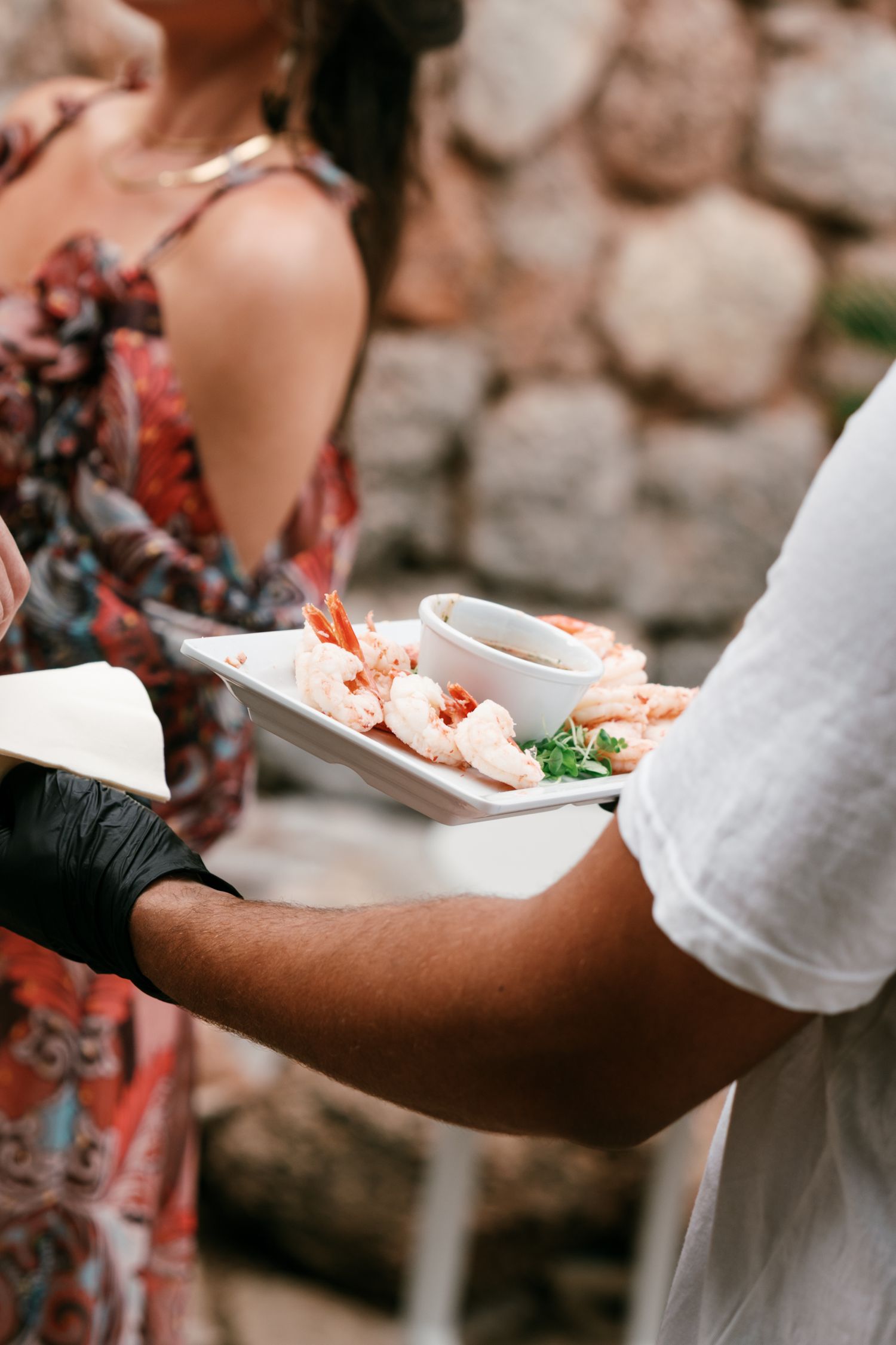 Server in black gloves presents a white tray of shrimp with dipping sauce and herbs to a guest at an outdoor stone-wall event.