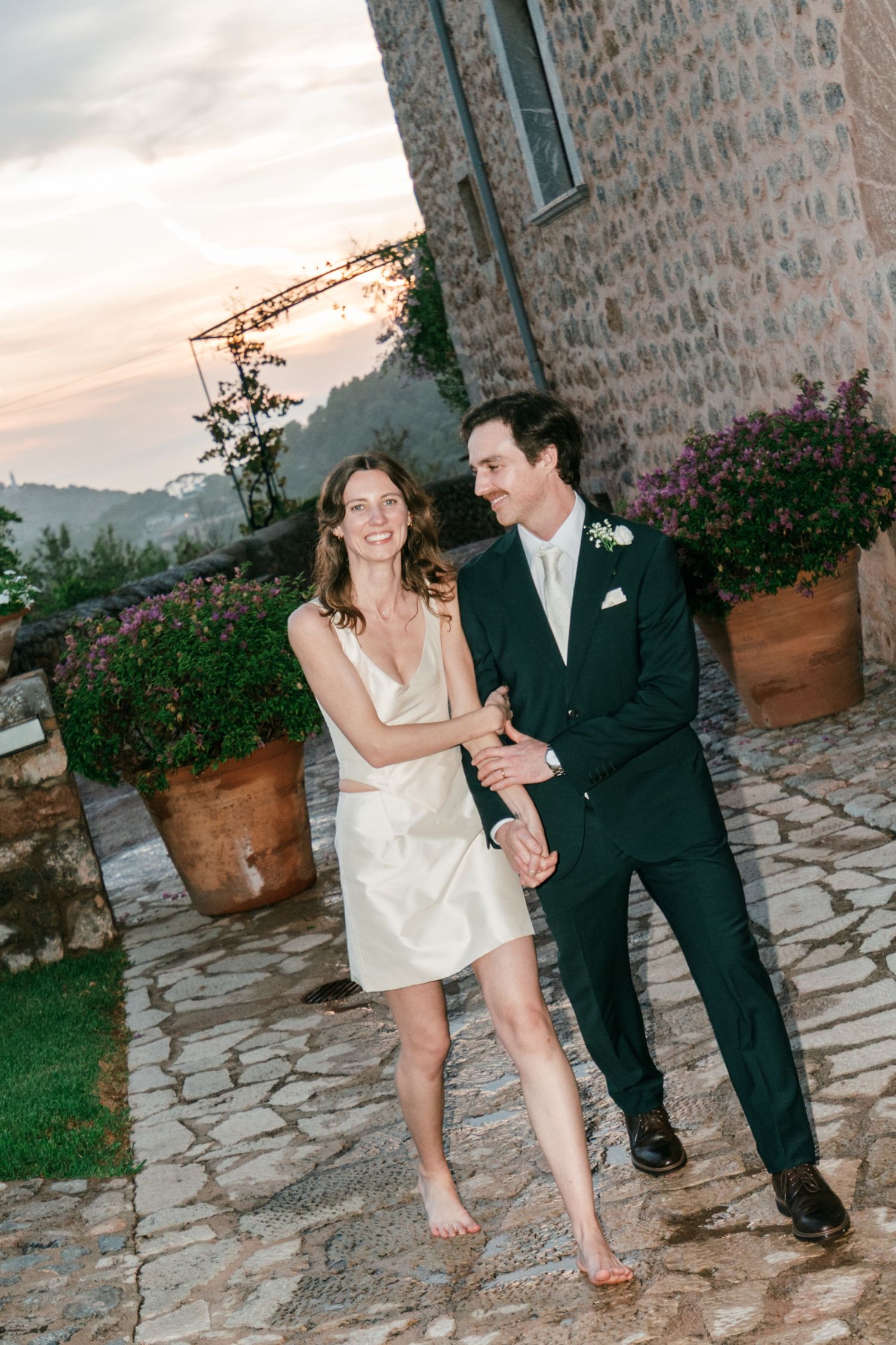 Barefoot bride in a short cream satin dress holds hands with a groom in a dark suit on a stone patio at sunset.