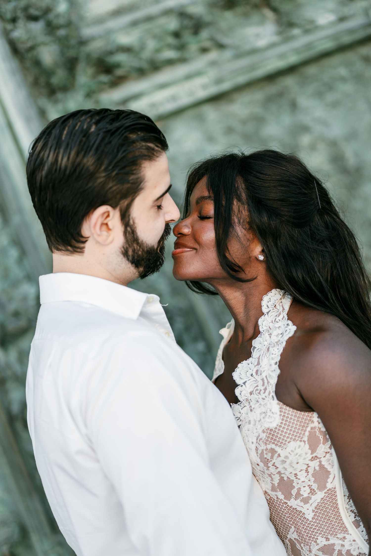 A bearded man in a white shirt and a woman in a lace dress lean in close for a romantic kiss during an outdoor couple portrait.