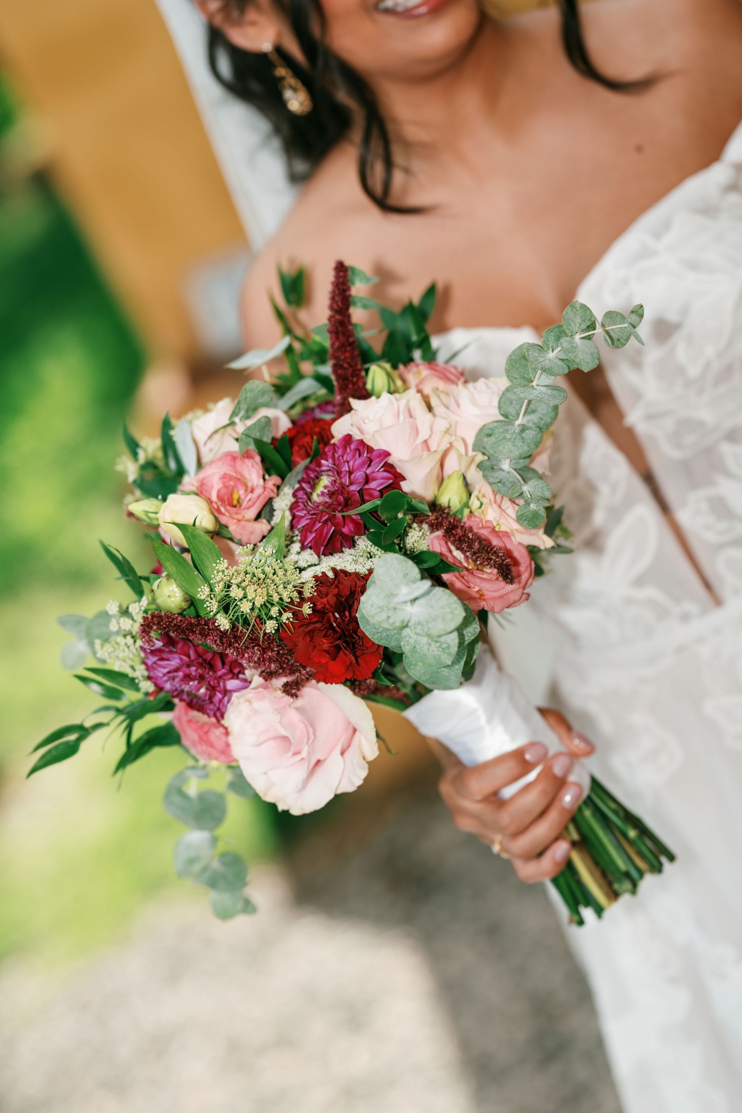 Bride in a white lace gown holding a wedding bouquet of pink roses, burgundy blooms, and eucalyptus leaves wrapped in a white ribbon.