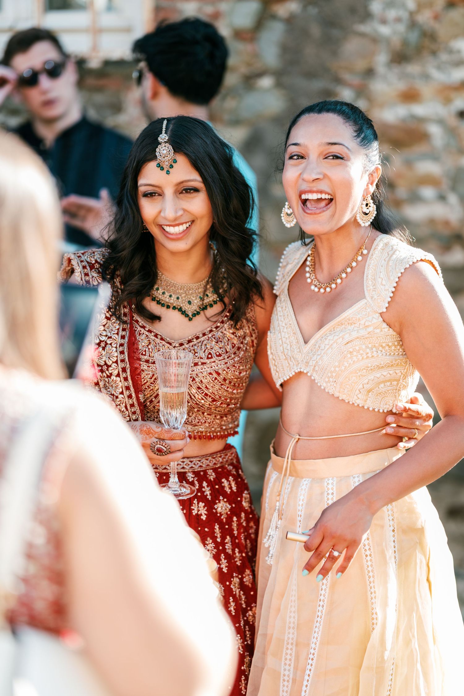 Two laughing women in embroidered red and cream lehengas with jewelry embrace at an outdoor Indian wedding celebration.
