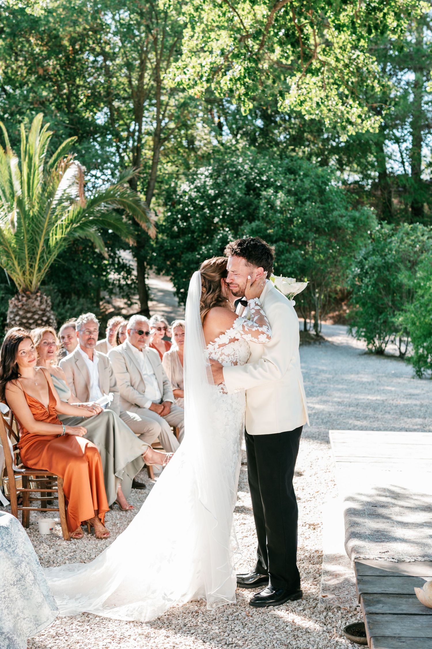 Bride and groom share first kiss at outdoor wedding ceremony with guests seated on wooden chairs among lush trees.