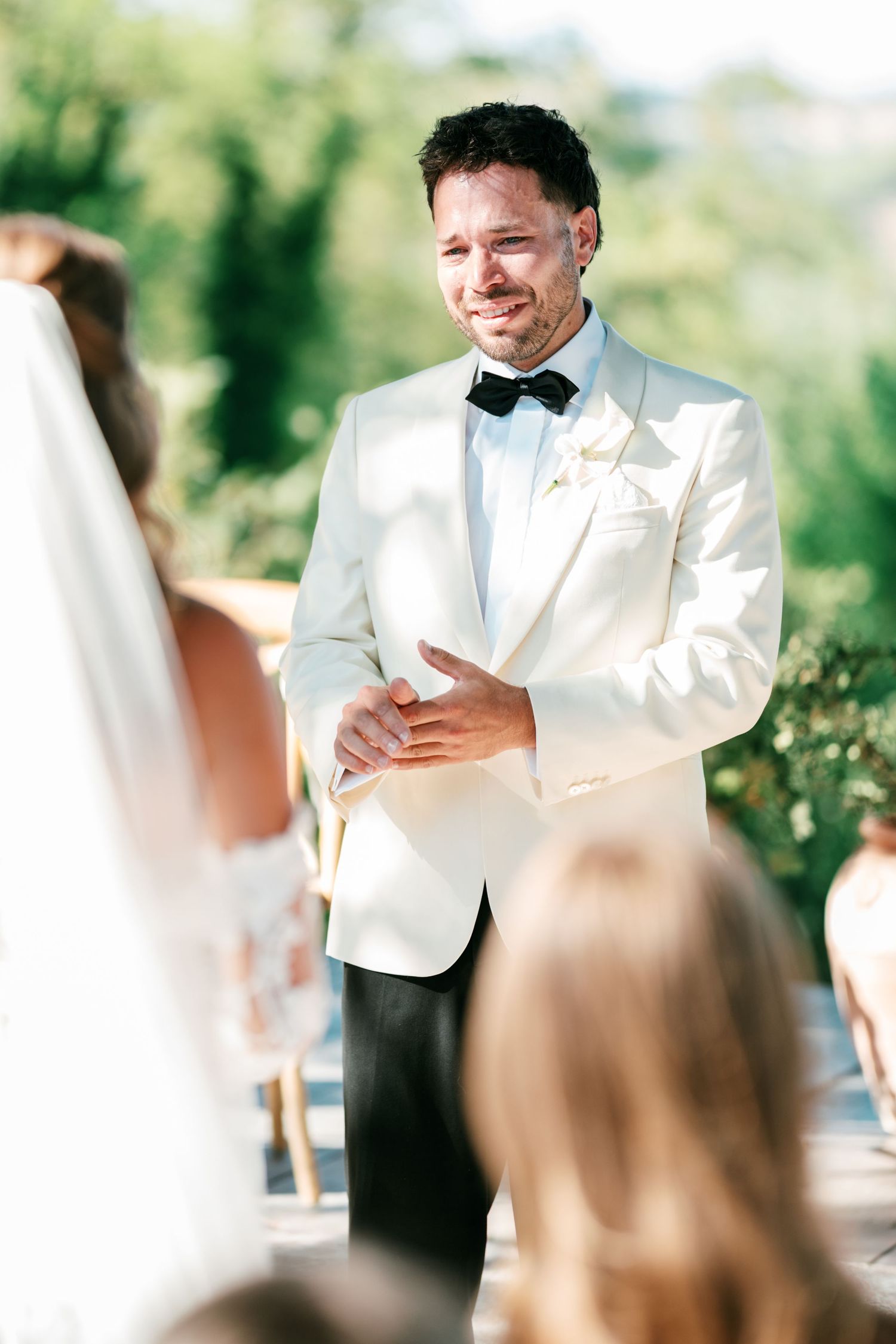 Emotional groom in white tuxedo jacket and bow tie reacts as bride walks down the aisle at outdoor wedding ceremony.