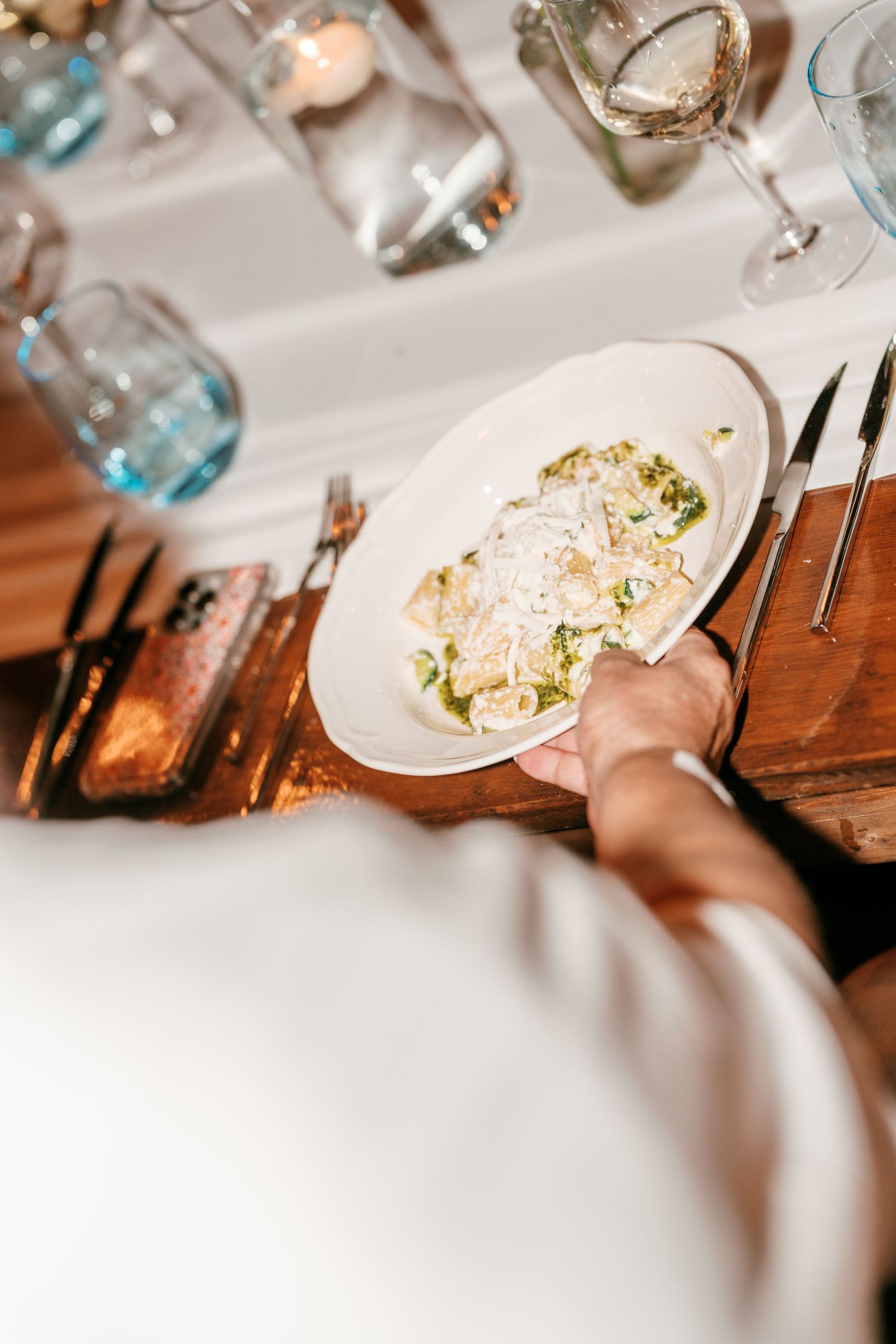 Server placing a white oval plate of pasta with pesto and parmesan on an elegant dining table with glassware.