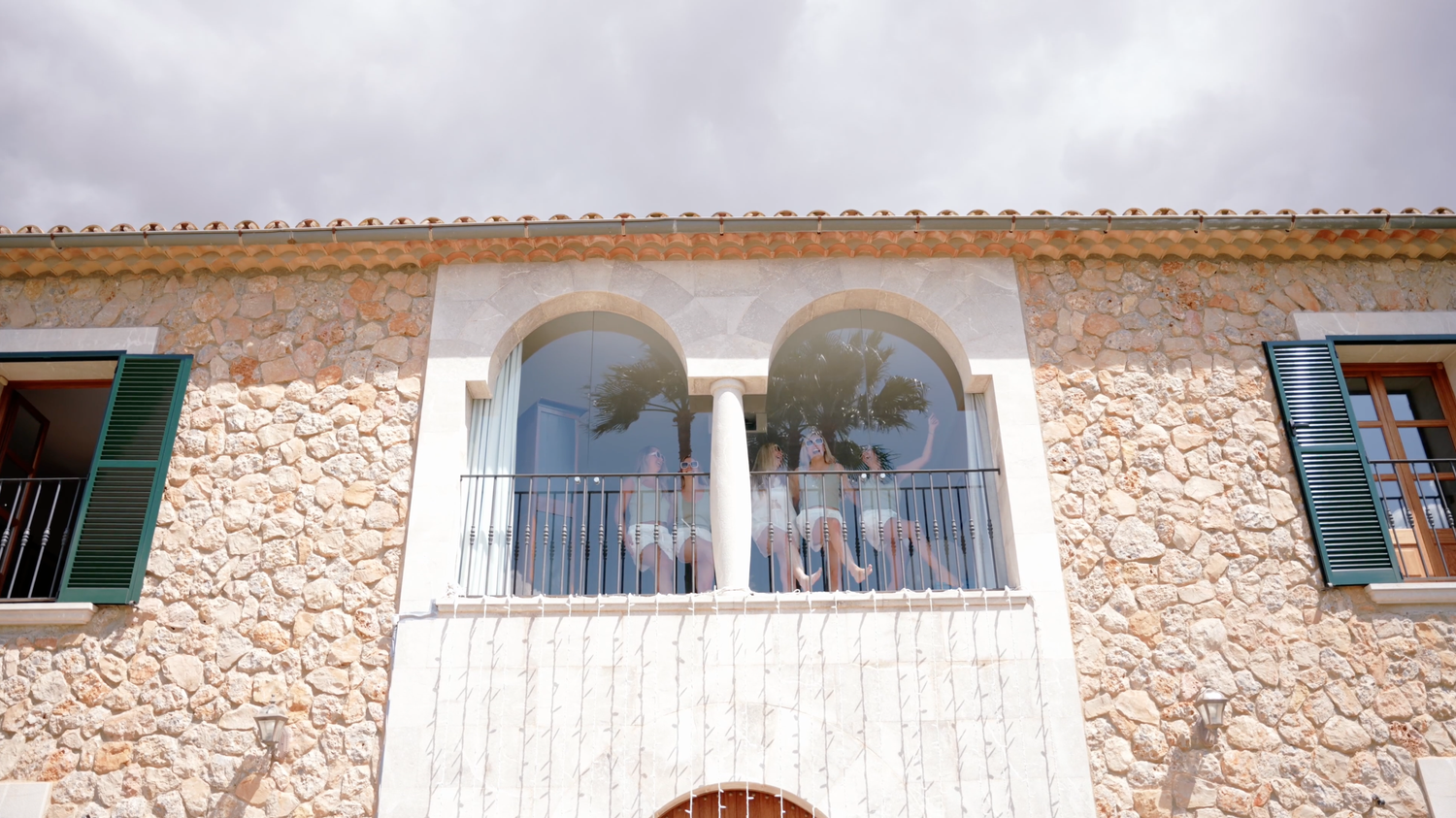 Rustic stone villa facade with a white arched balcony, green wooden shutters and guests gathered on the terrace under a cloudy sky.