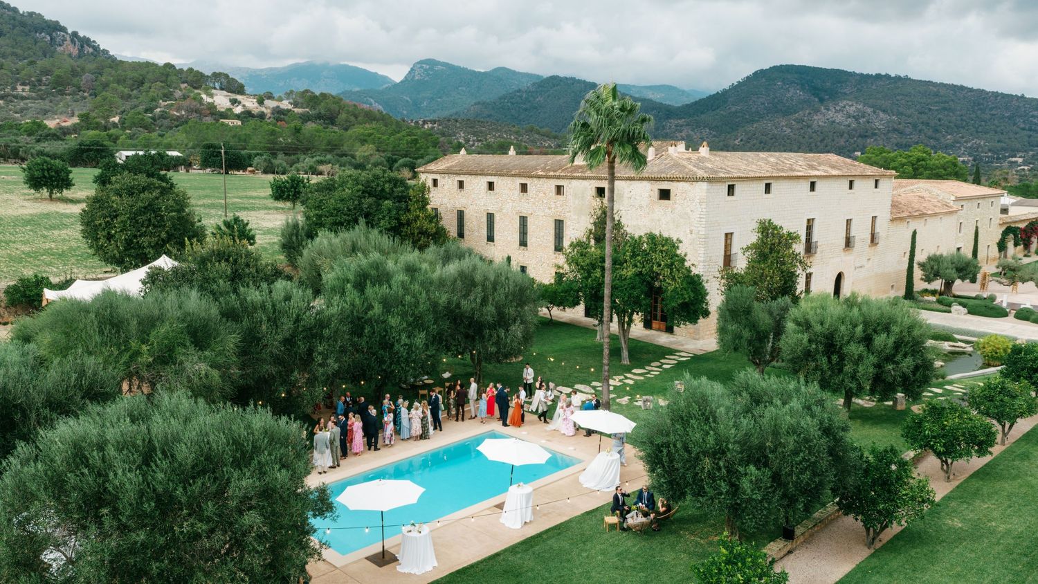 Wedding guests gather beside a turquoise pool at a stone countryside villa surrounded by olive trees, palms and mountain views.