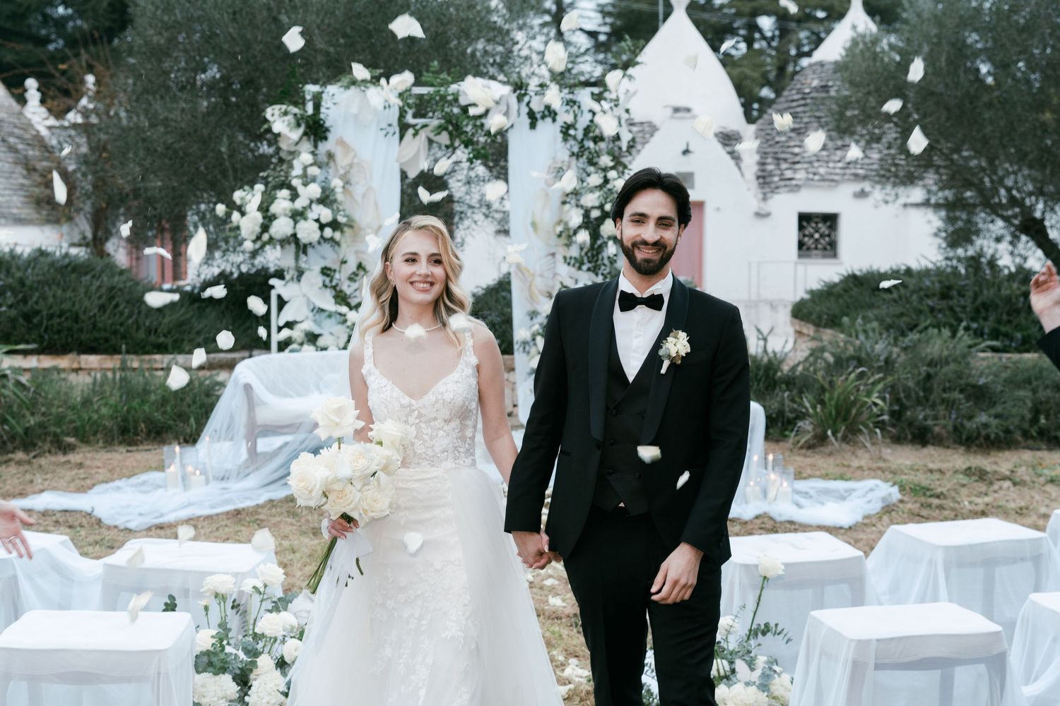 Smiling newlyweds walk hand in hand through flower petal confetti at outdoor wedding ceremony. Puglia wedding photographer, film
