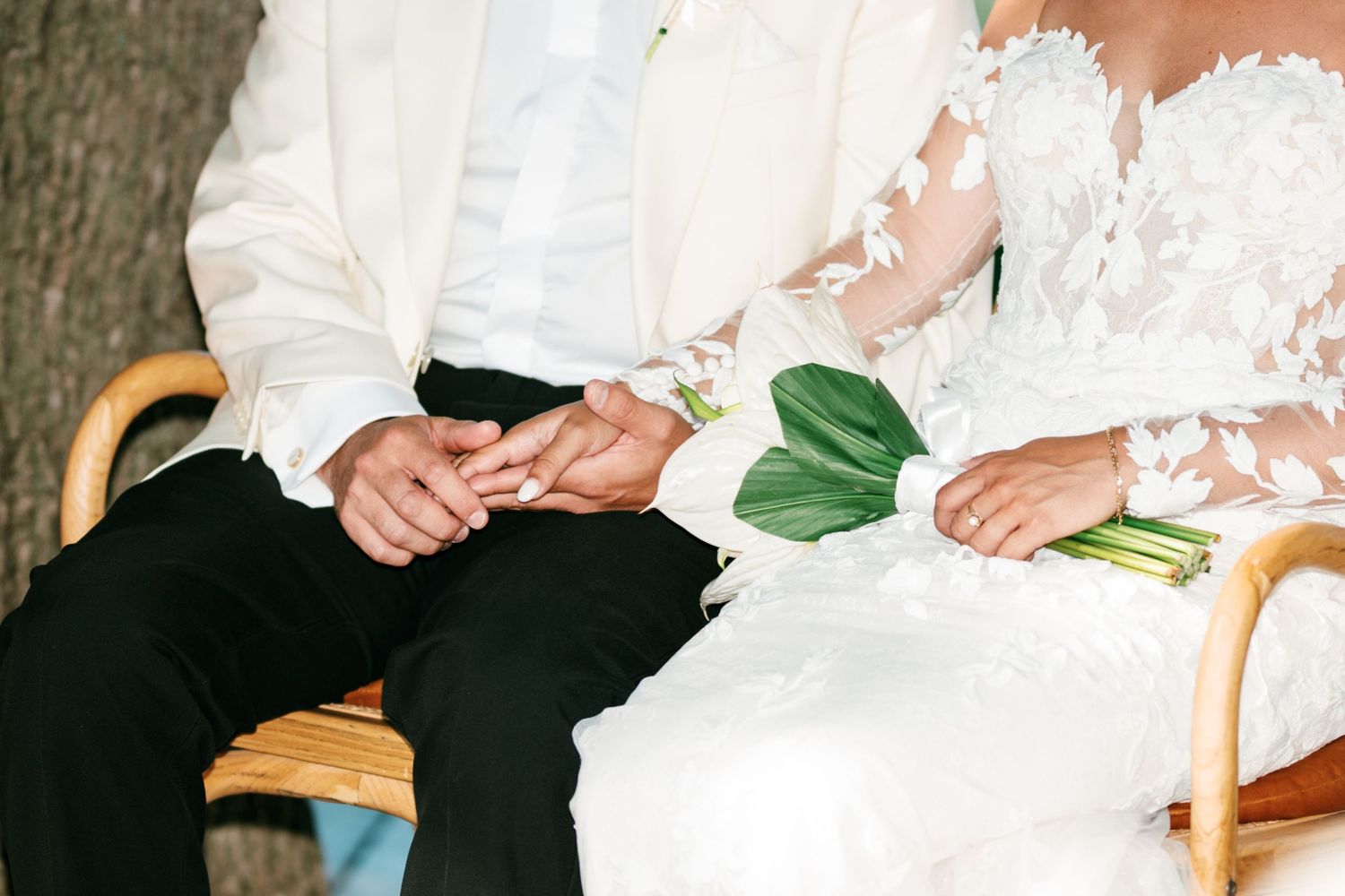 Bride and groom holding hands while seated, bride holding green bouquet, wearing white lace gown and groom in white suit.