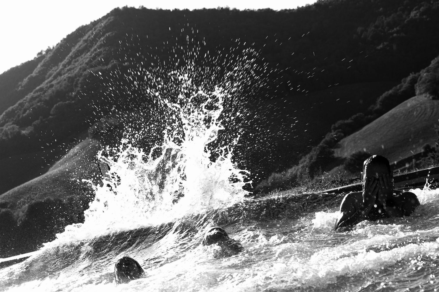 Black and white photo of powerful ocean waves crashing against rocky shore with green hillside in background.