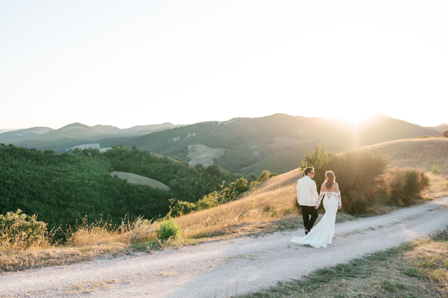 A bride and groom walk hand in hand on a dirt path with golden sunset over rolling green hills.