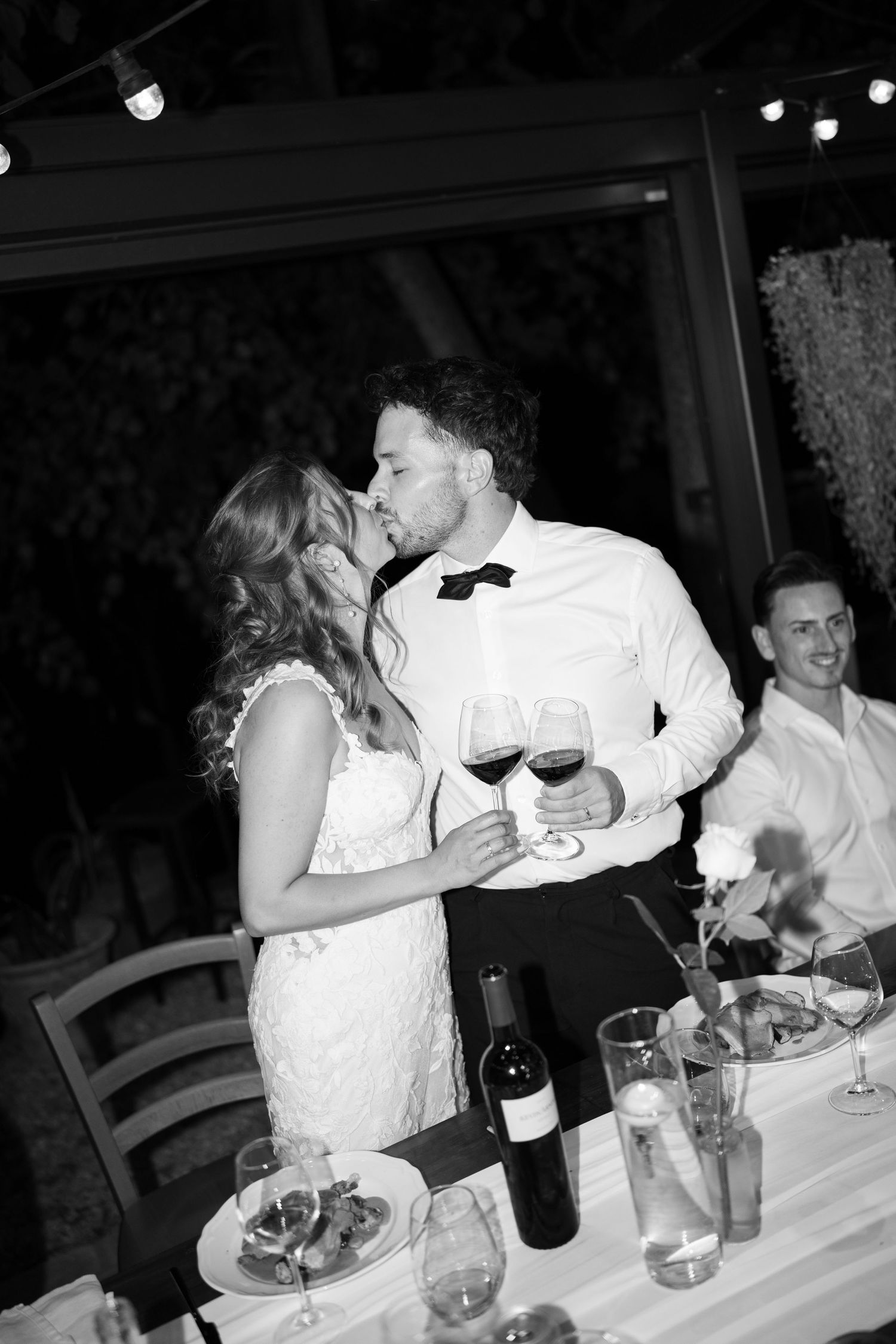 Black and white photo of a bride and groom sharing a kiss at their wedding reception dinner table with wine glasses.