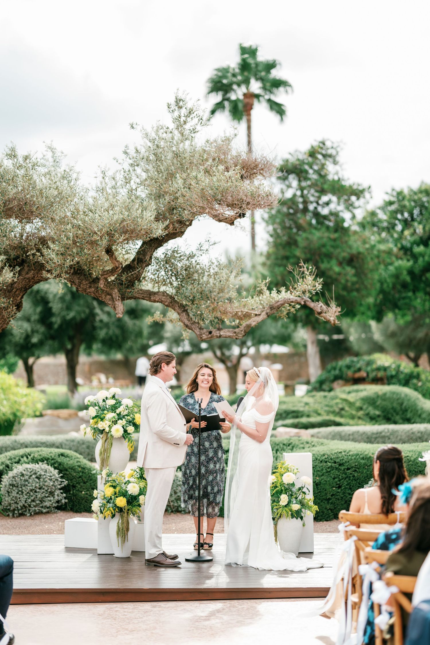 Bride and groom exchange vows during an outdoor garden wedding ceremony under an olive tree as guests watch from wooden chairs.