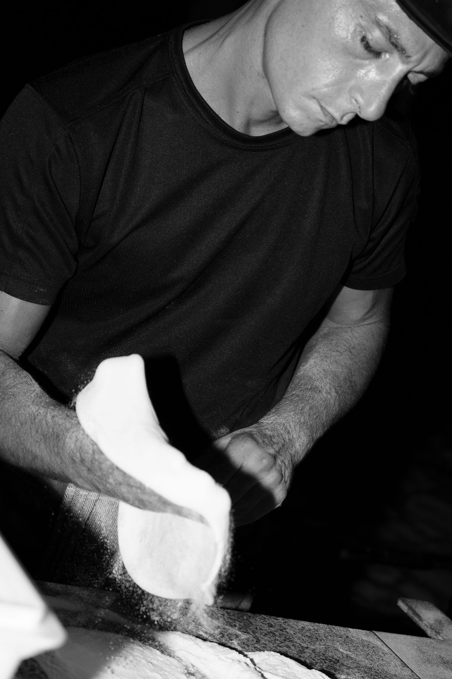 Black and white photo of a pizza maker in a dark t-shirt stretching fresh dough with flour dusting the air.