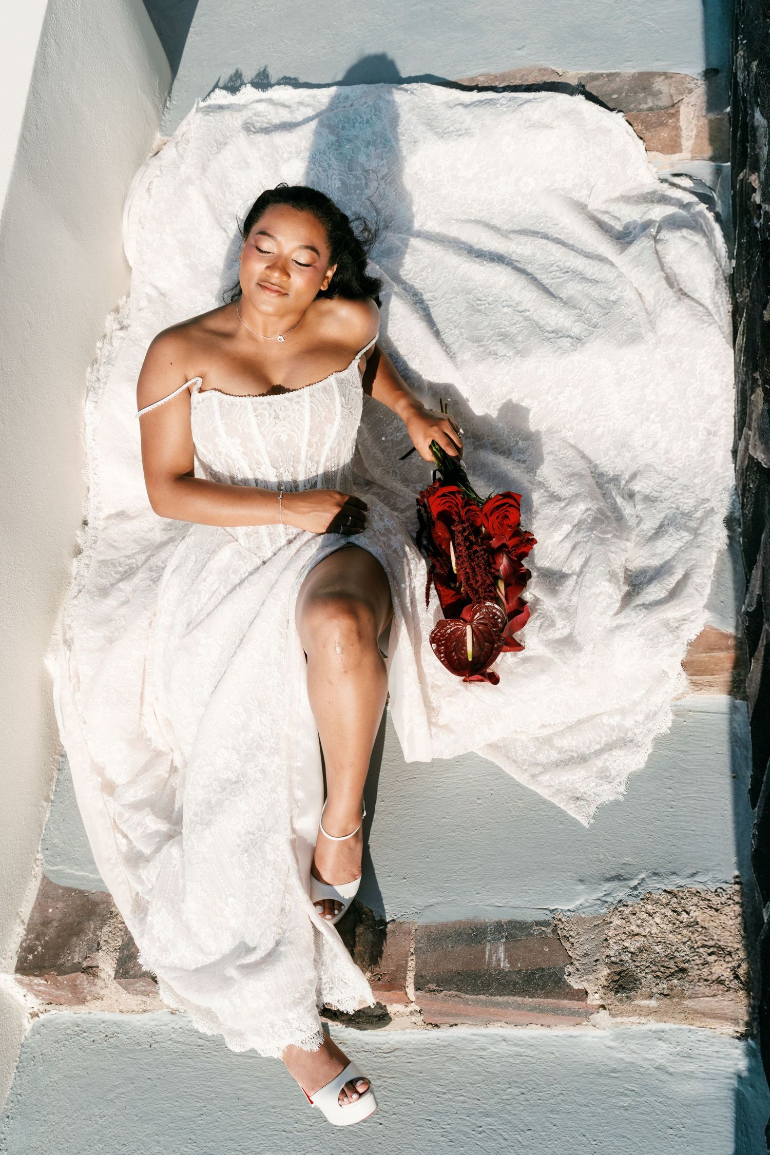 Bride in white dress relaxes on sunlit Santorini steps holding red heeled sandals, eyes closed peacefully.