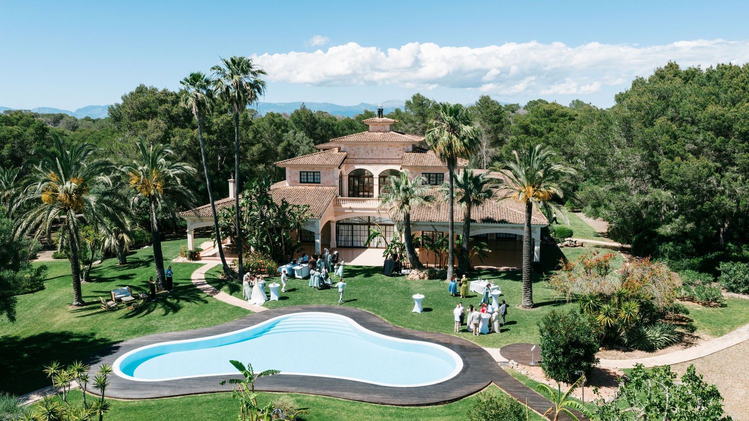 Aerial view of a luxurious Mediterranean villa with a pool, lush palm trees, and an elegant outdoor wedding setup.
