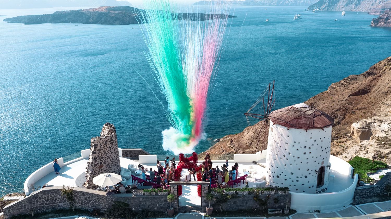 Colorful fireworks display over a scenic Santorini cliffside windmill with crowd gathered, overlooking the stunning blue Aegean Sea.