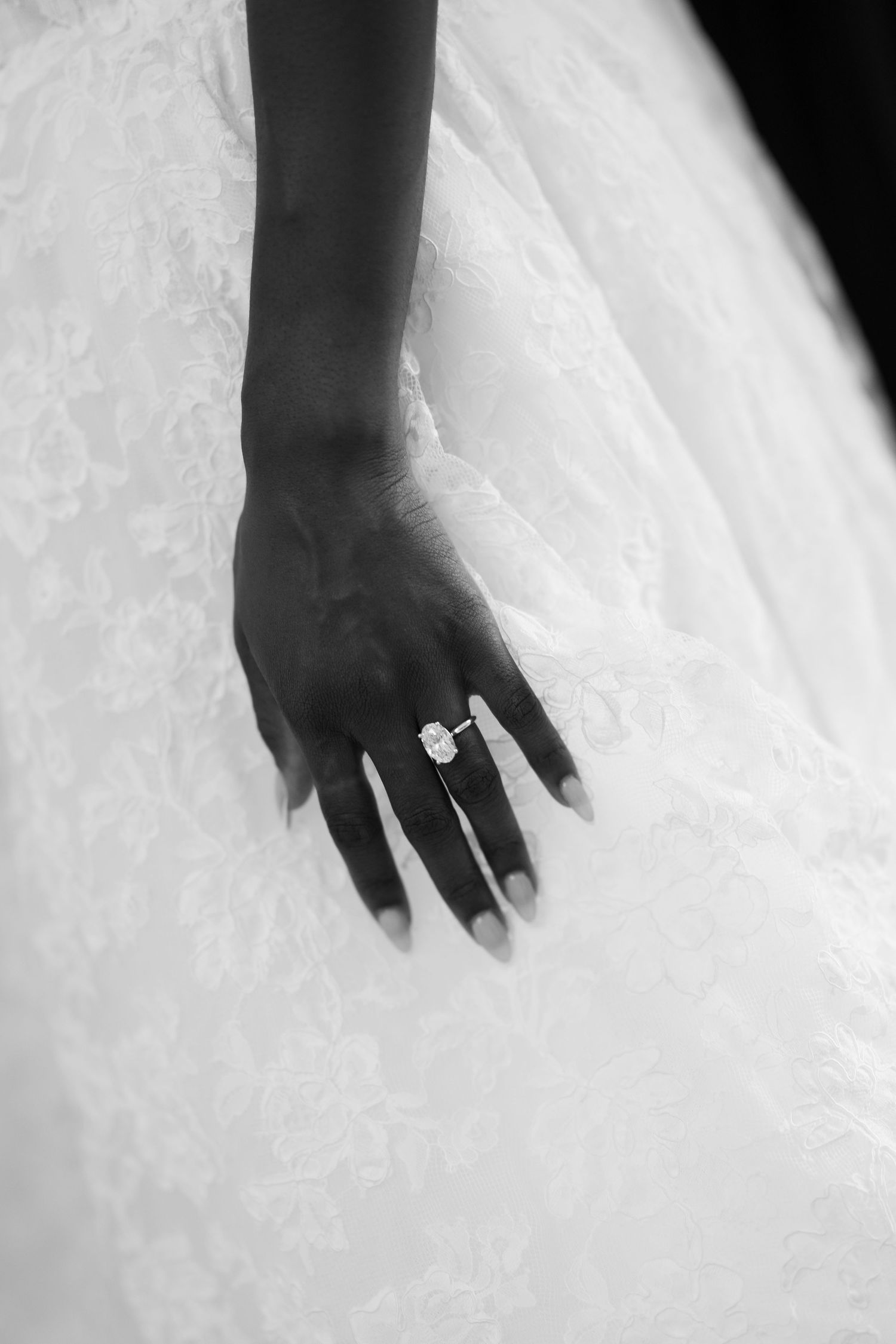 Black and white close-up of a bride's hand with a sparkling diamond engagement ring resting on a lace wedding gown.