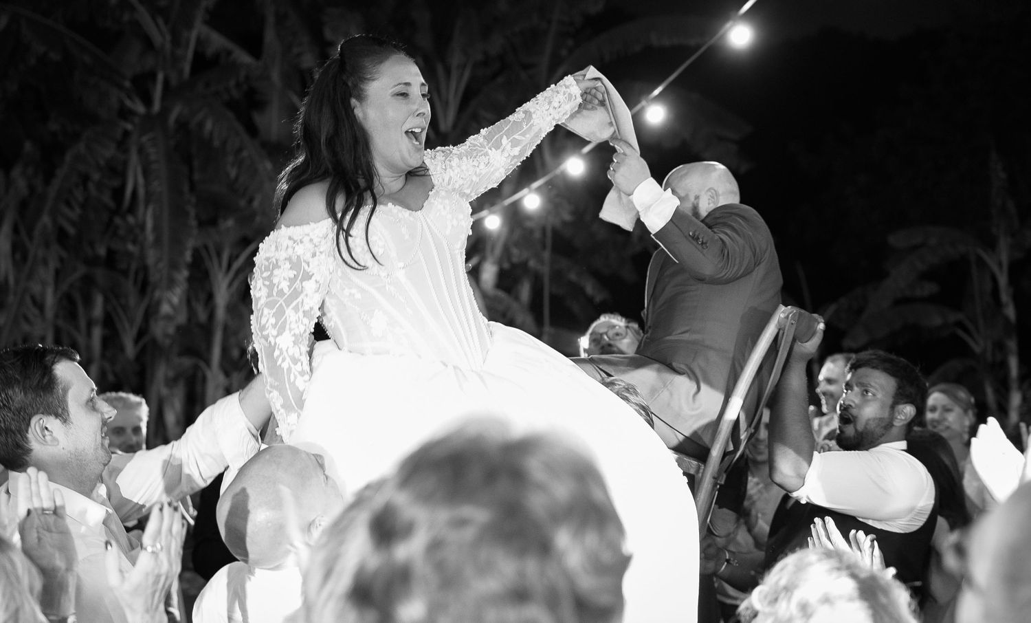 Bride in a lace wedding gown lifted in a chair above cheering guests during a joyful hora dance at a nighttime outdoor reception.