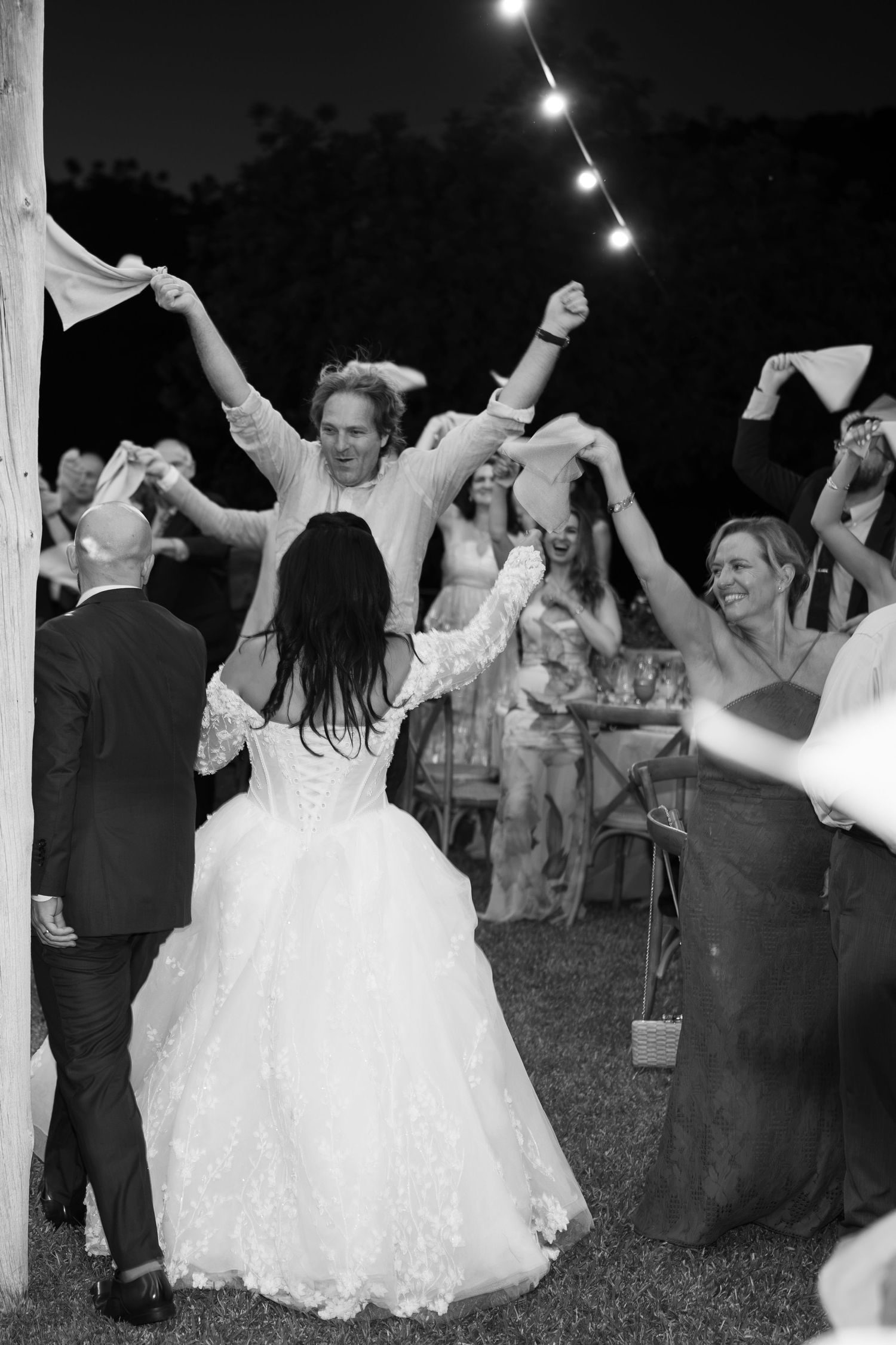 Bride in a lace wedding gown dances outdoors under string lights as guests wave white napkins in a lively black and white celebration.