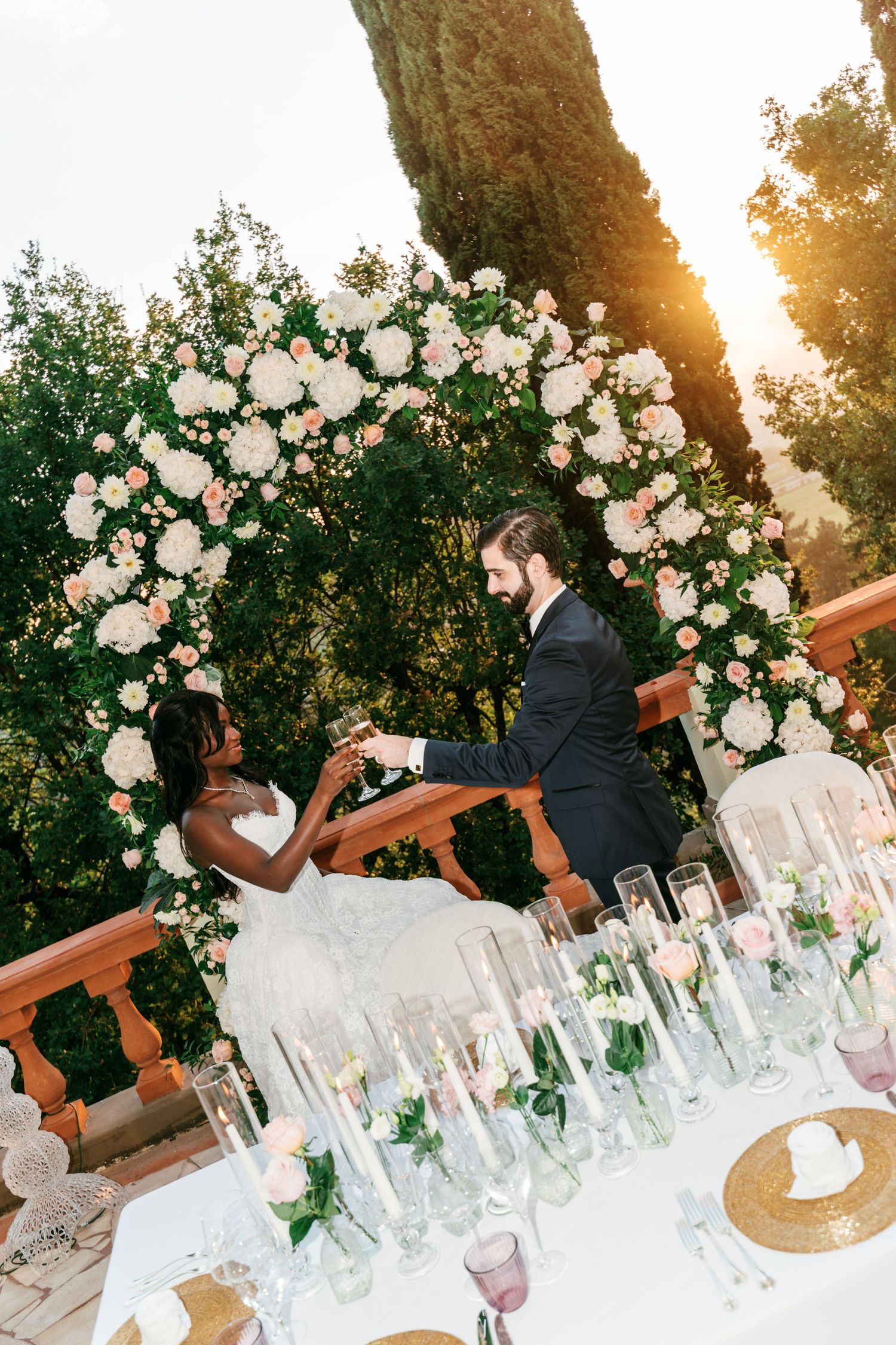 Bride in a white gown and groom in a black tuxedo toast with champagne beneath a floral wedding arch at their outdoor reception table.