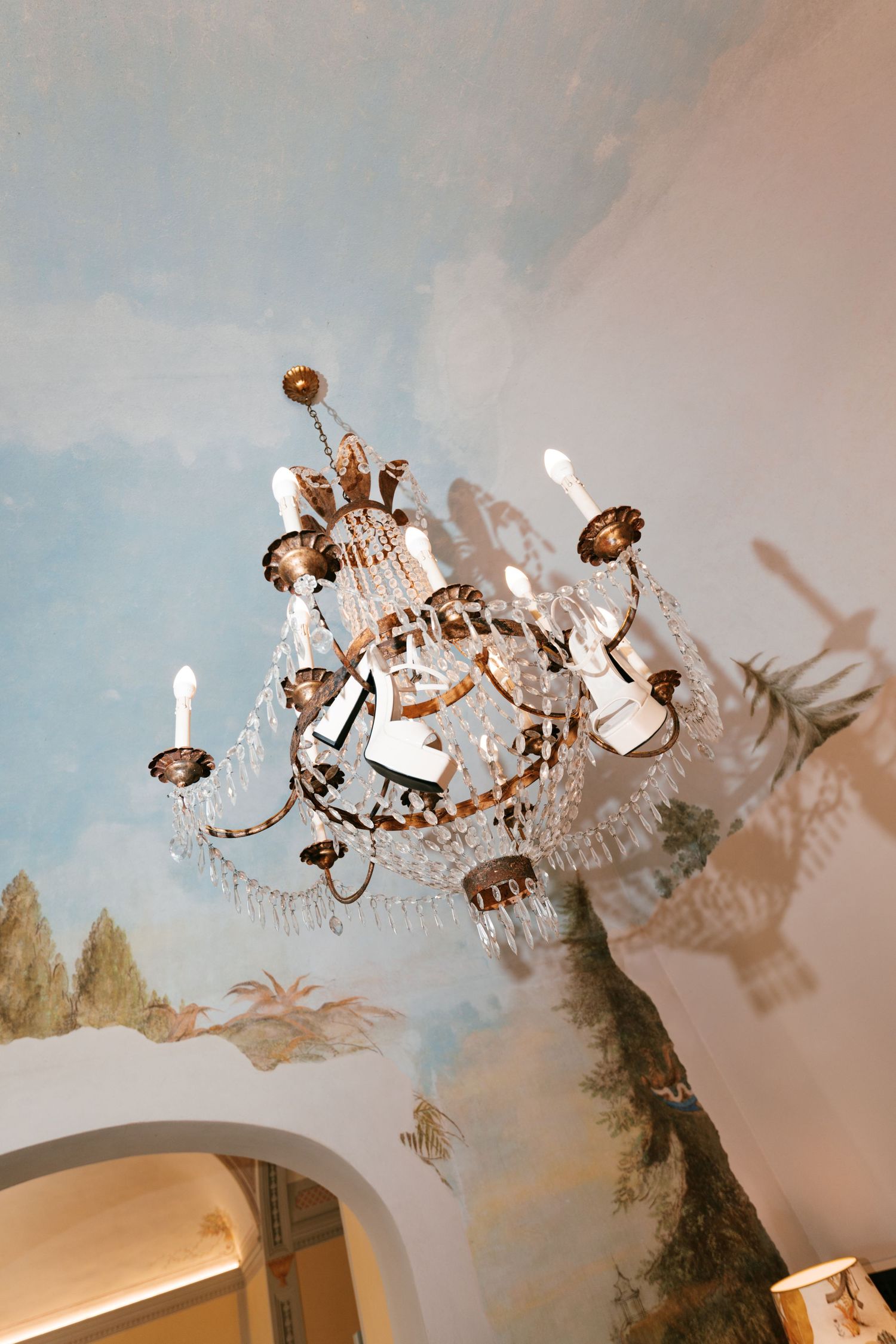 Ornate crystal chandelier with candle-style lights hangs beneath a ceiling painted with a soft blue sky, clouds and trees.