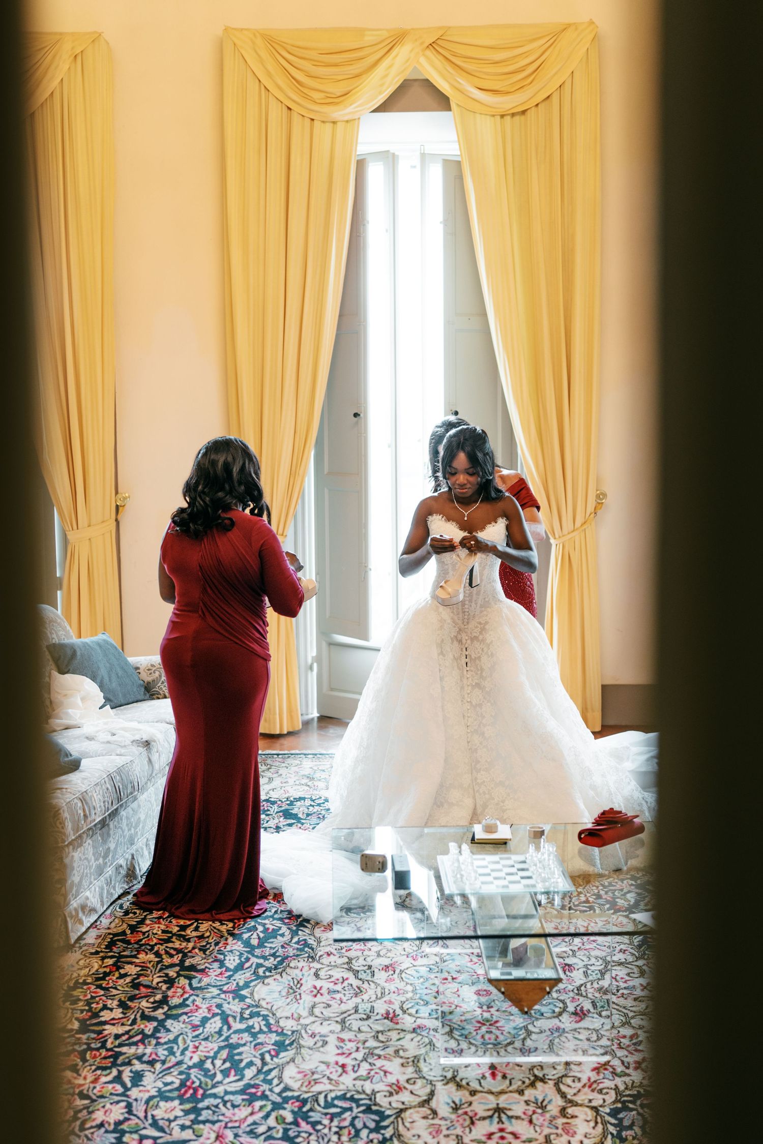 Bride in a white strapless ball gown stands before a mirror in an elegant room while a woman in a red dress helps her prepare.