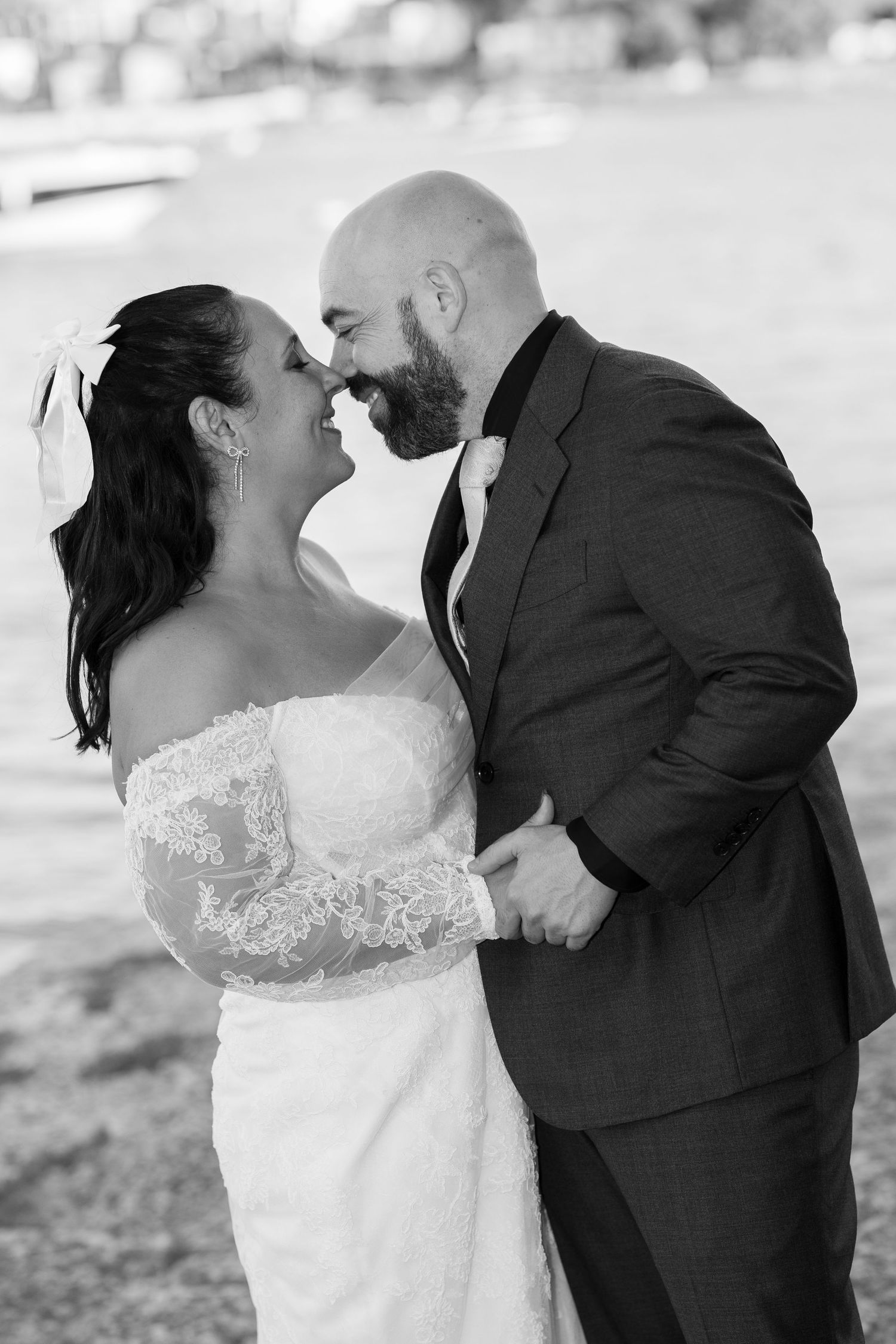 Black and white photo of a smiling bride in a lace off-shoulder gown and a bearded groom in a suit touching noses by the water.