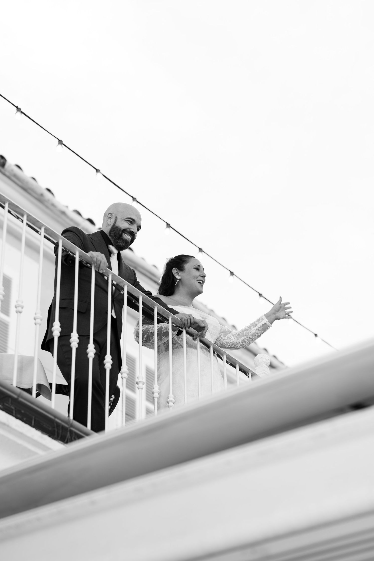 Black and white photo of a smiling couple on a balcony with a white railing, the bride gesturing outward, string lights overhead.