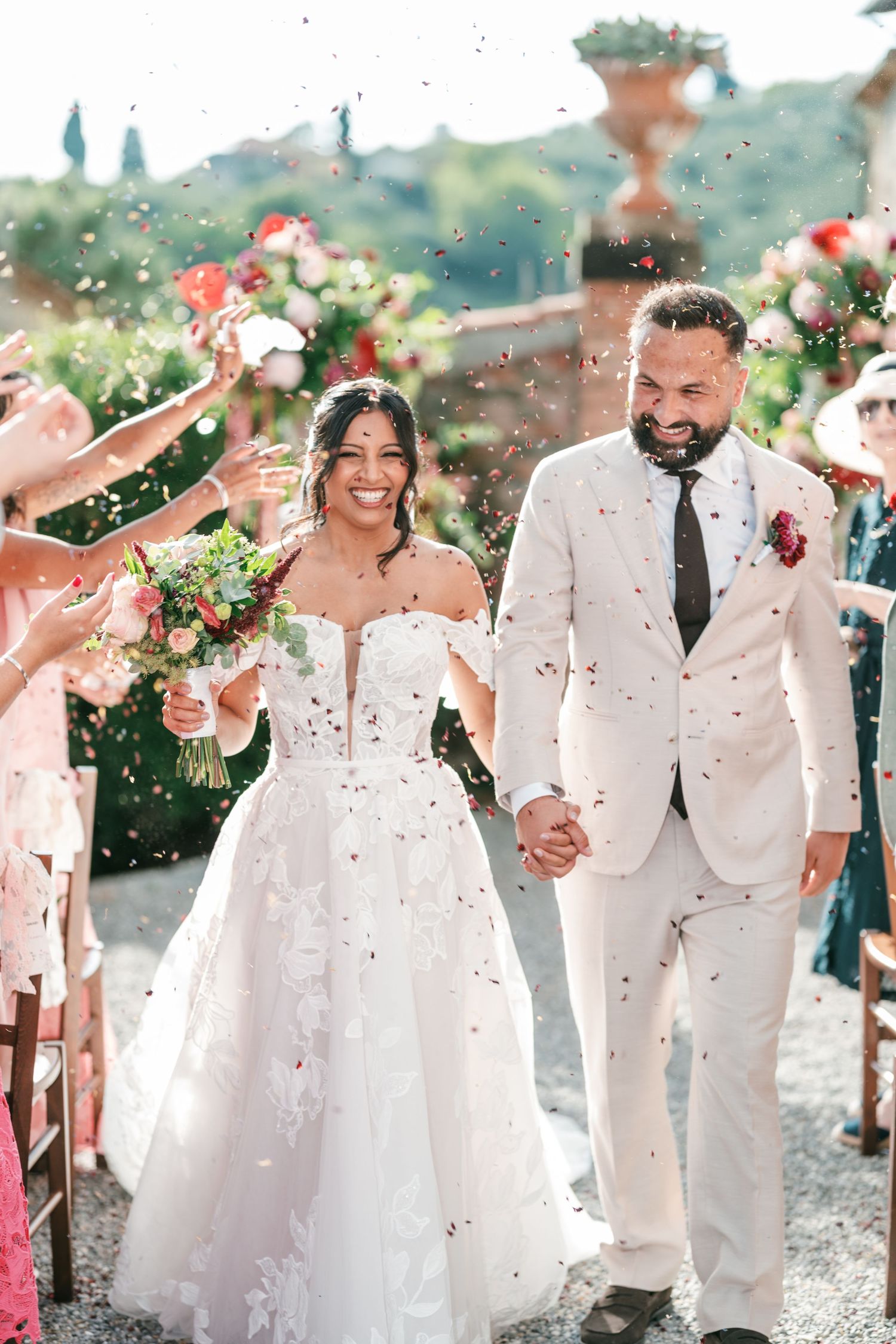 Smiling bride in a lace wedding gown and groom in a cream suit walk hand in hand as guests toss confetti at an outdoor garden wedding.