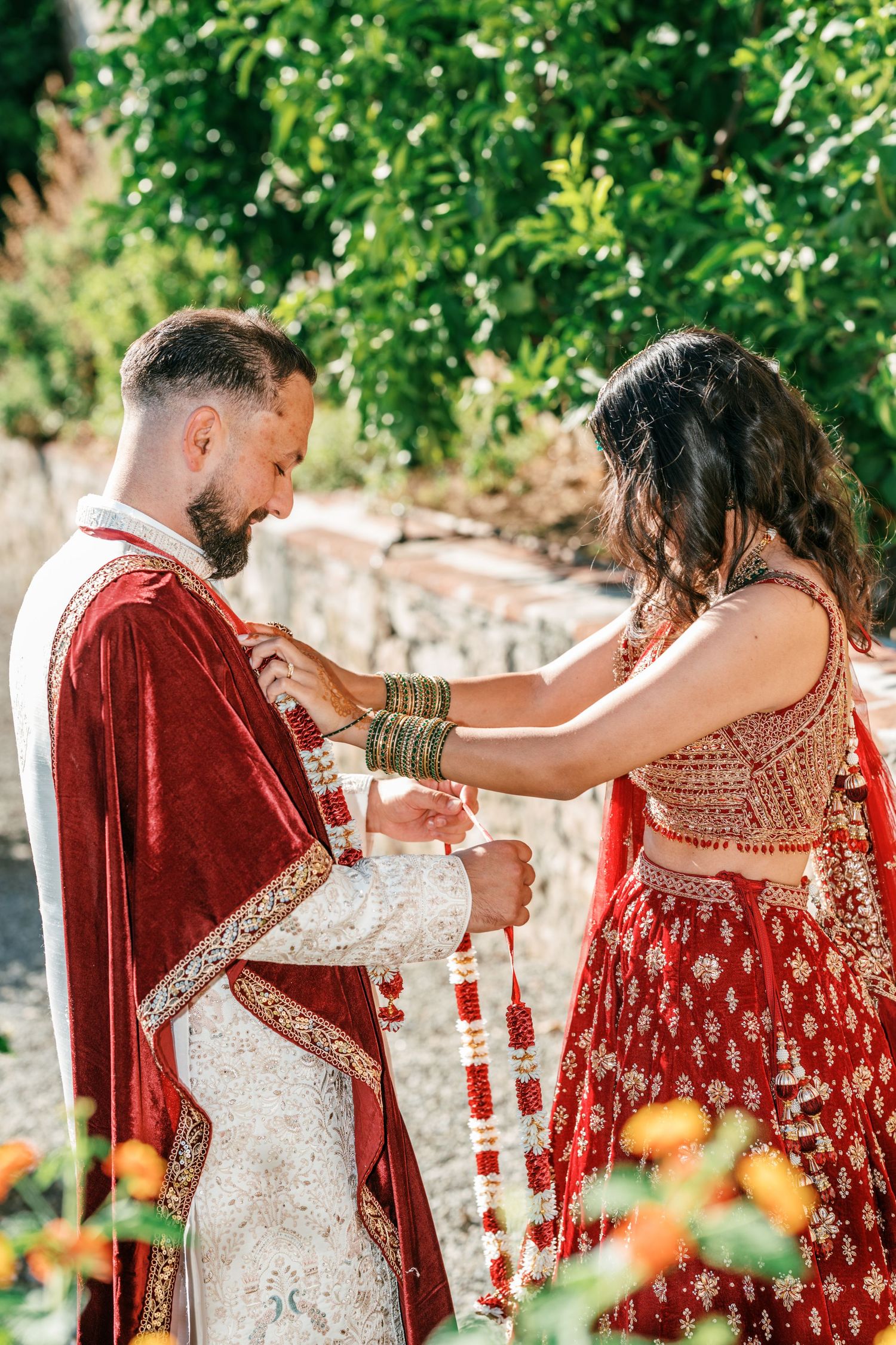 Groom in a red velvet cape and bride in an embroidered red lehenga exchange floral garlands in a traditional Indian wedding ceremony.