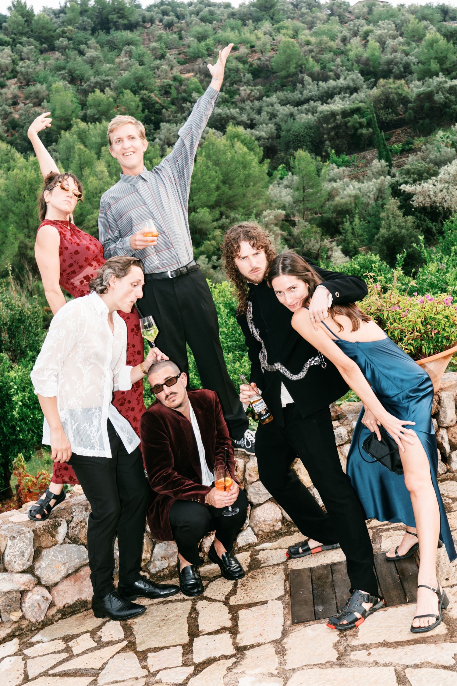 Six stylishly dressed friends pose playfully with drinks on a stone terrace, with lush green hillside and trees in the background.