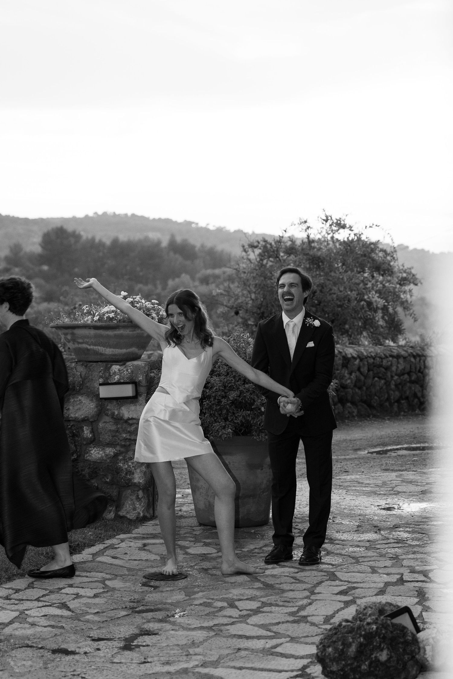Black and white photo of a barefoot bride in a short satin dress dancing joyfully with a laughing groom in a suit on a stone terrace.