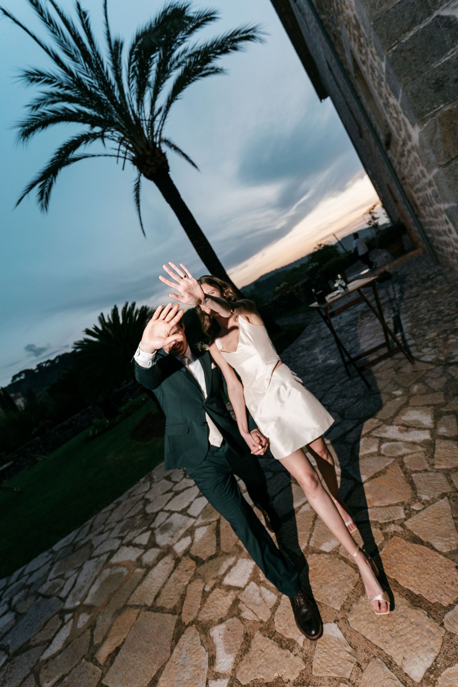 A wedding couple covers their faces with their hands on a stone patio beneath a palm tree and a dramatic evening sky.